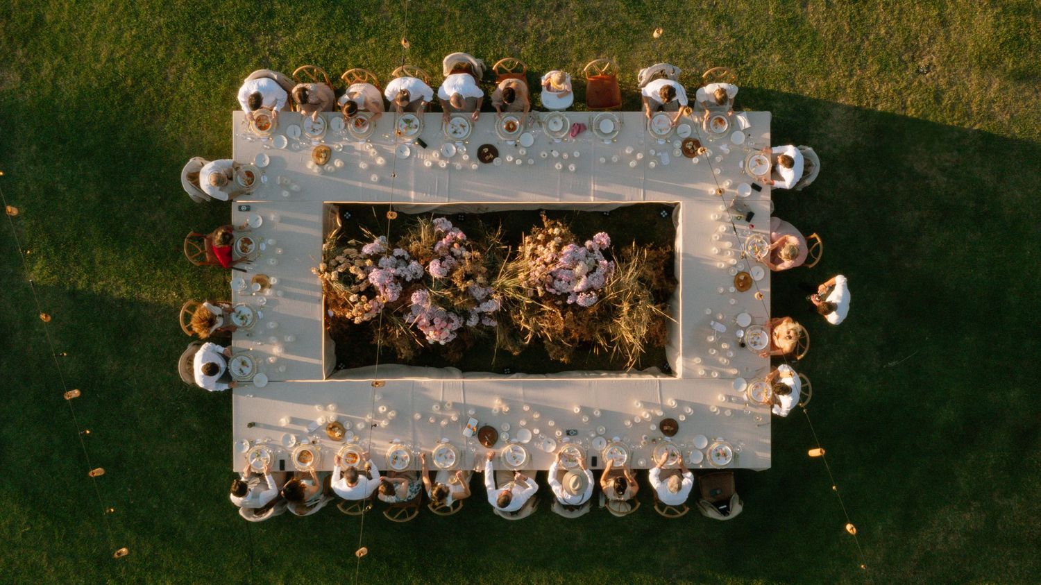 Aerial view of a wedding party arranged around a rectangular frame on green grass with string lights.