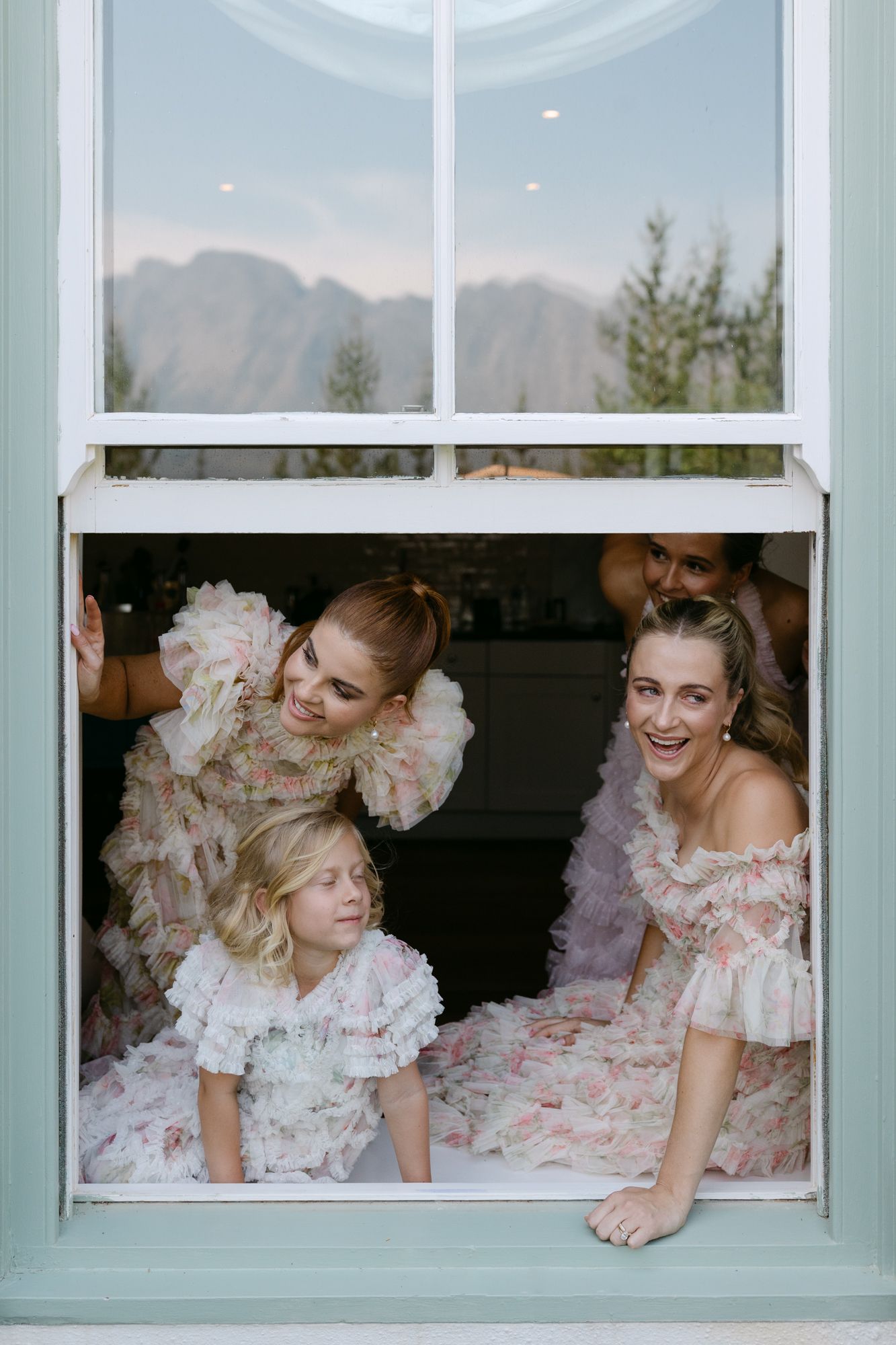 Three bridesmaids in blush pink dresses peek playfully through a window frame at a wedding venue.