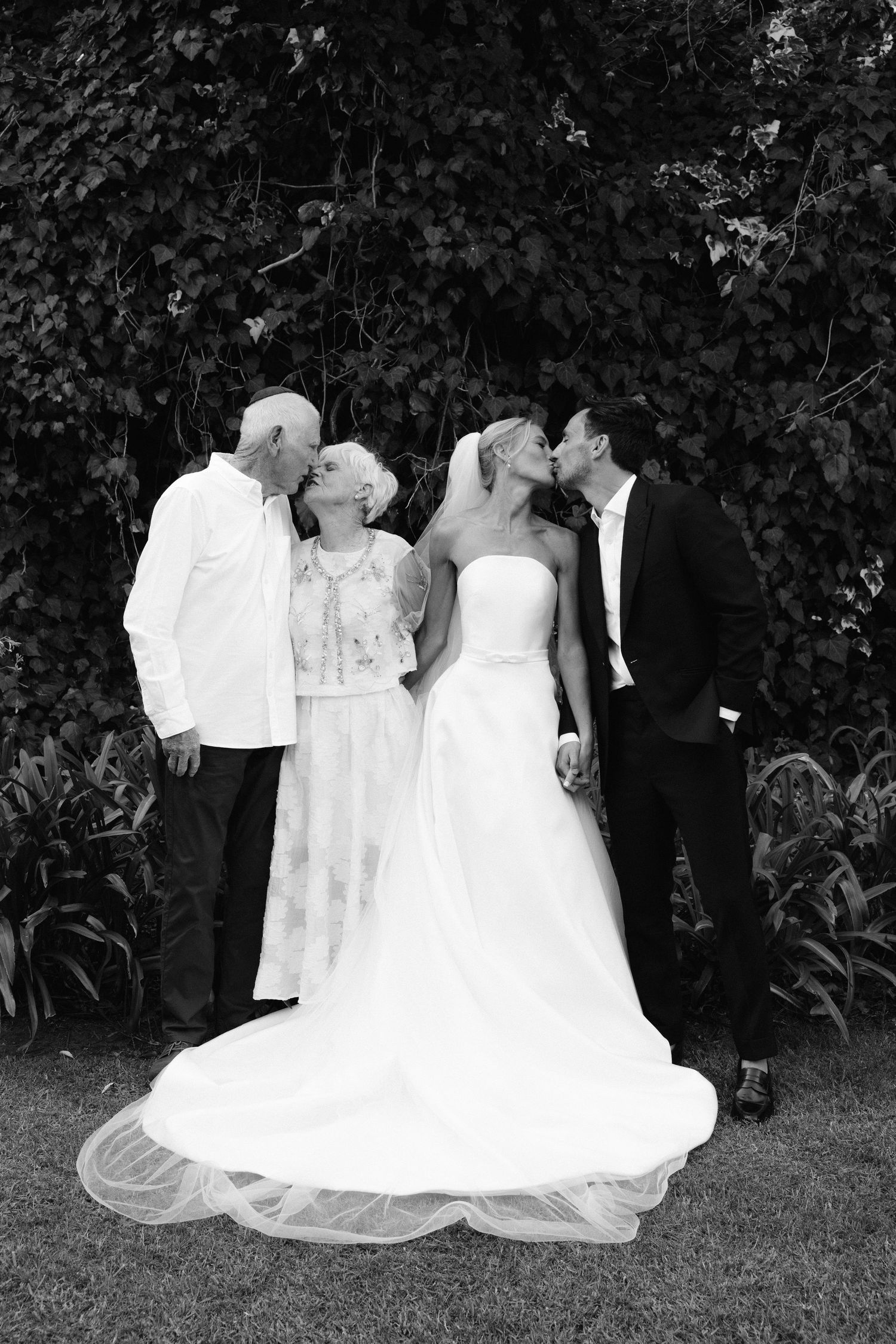 A black and white wedding photo showing two couples sharing an intimate moment in a garden setting.