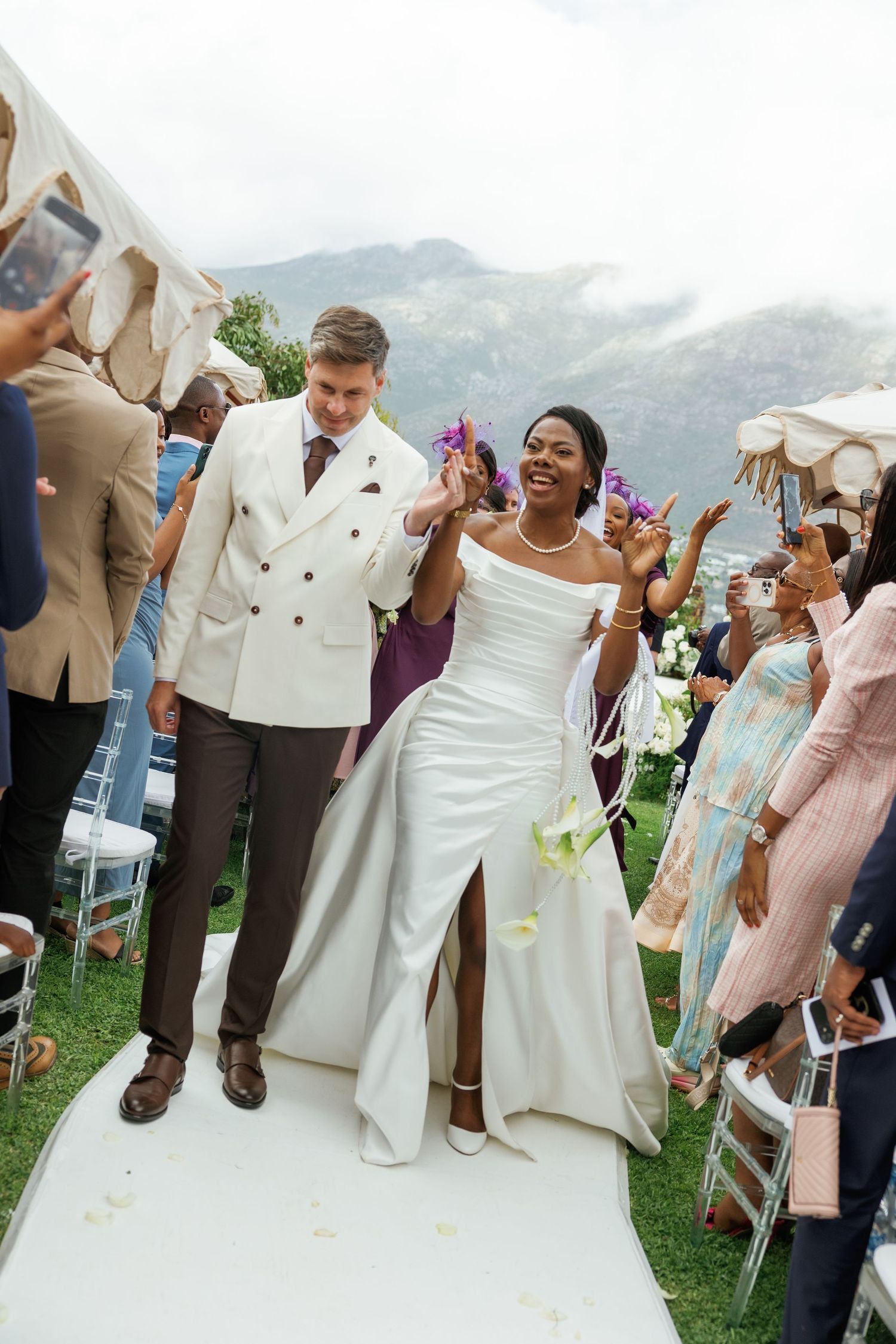 Interracial couple walks down wedding aisle outdoors, bride in white gown, groom in cream blazer, mountains in background.