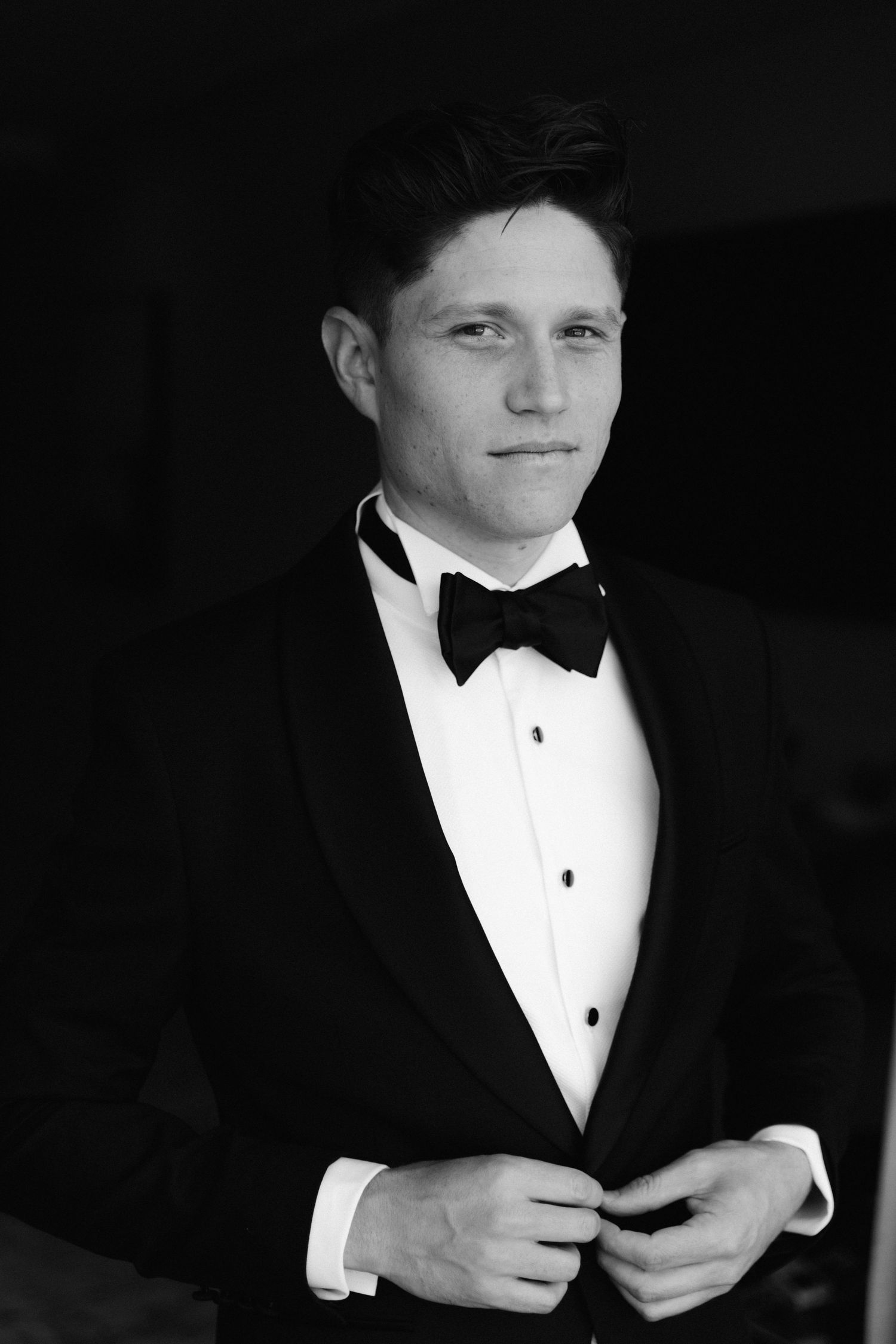 A person in a black tuxedo and white bowtie adjusts their cuffs in a dramatic black and white photograph.