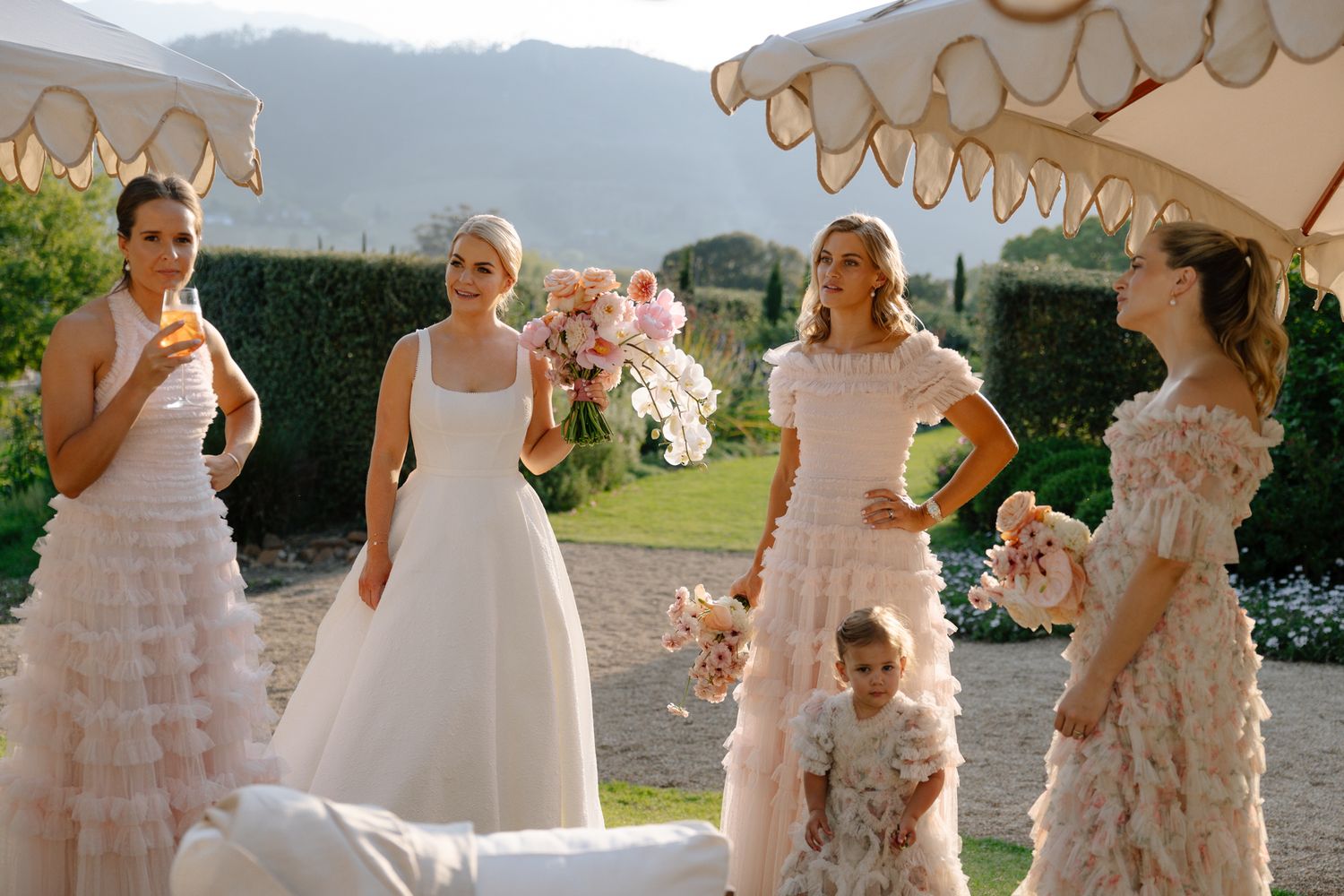 A group in white formal dresses stands outdoors with mountains in the background on a sunny day.