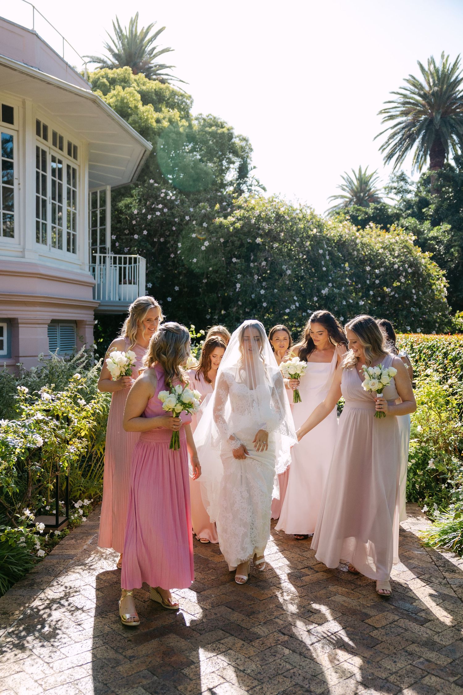 Bride in white lace gown and veil walks with bridesmaids in pink dresses holding bouquets in a garden.