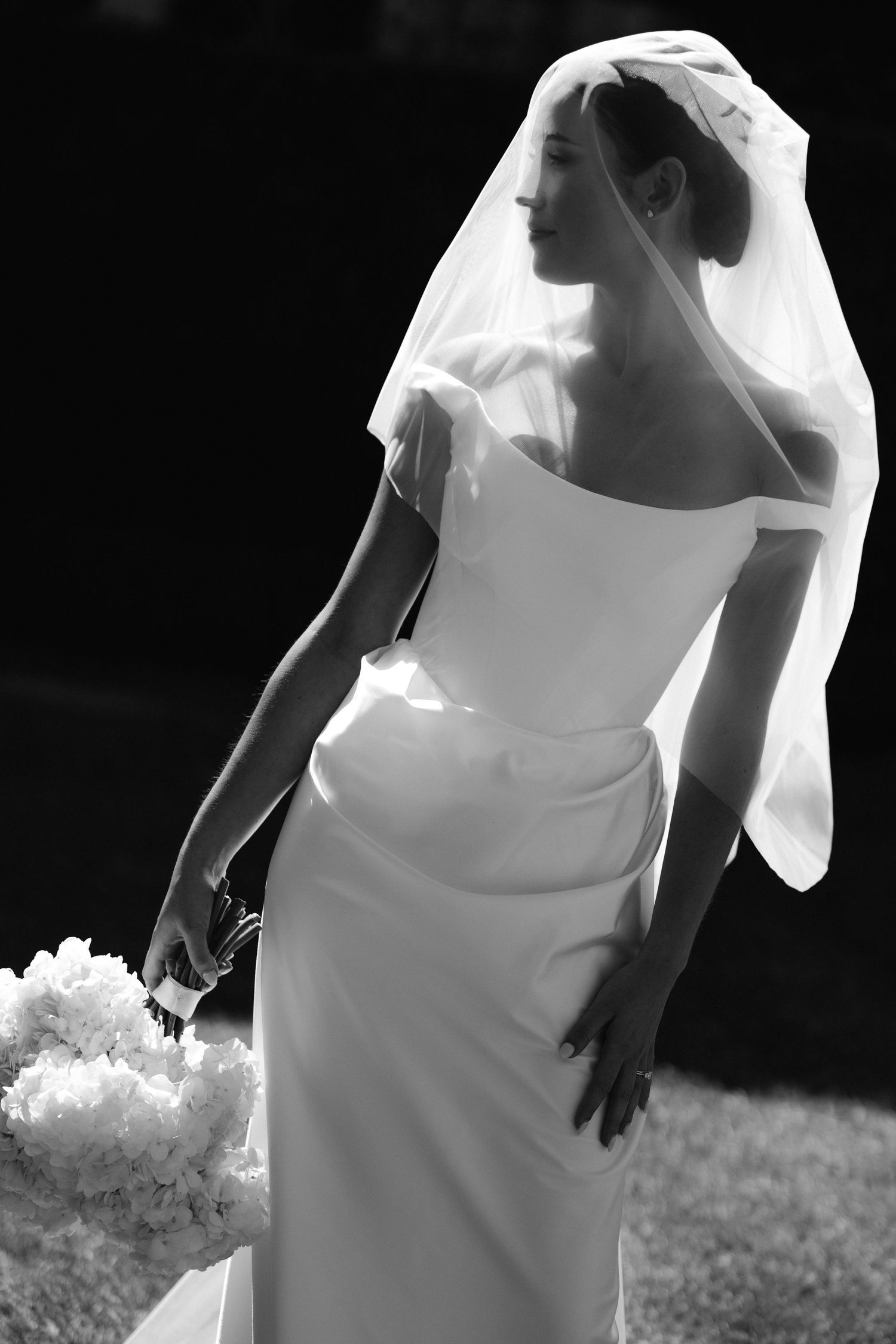Elegant bride in white off-shoulder gown and veil holds floral bouquet in black and white wedding portrait.