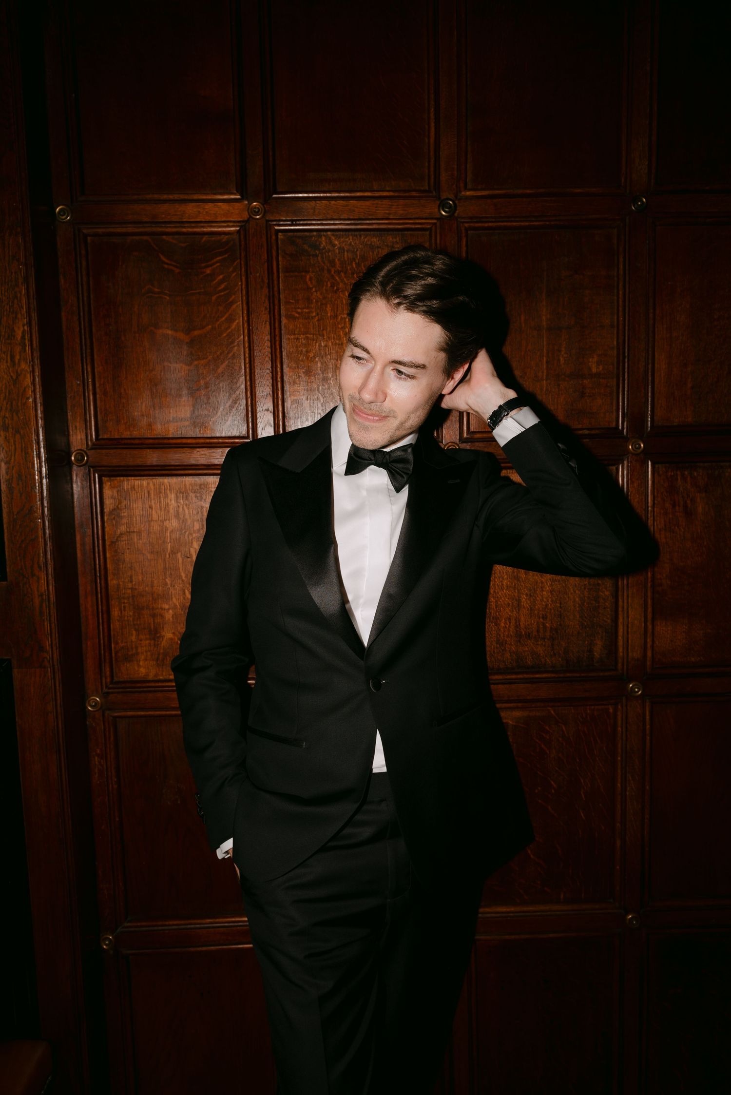 A person in a black tuxedo poses against a dark wood-paneled wall in a series of dramatic formal portraits.