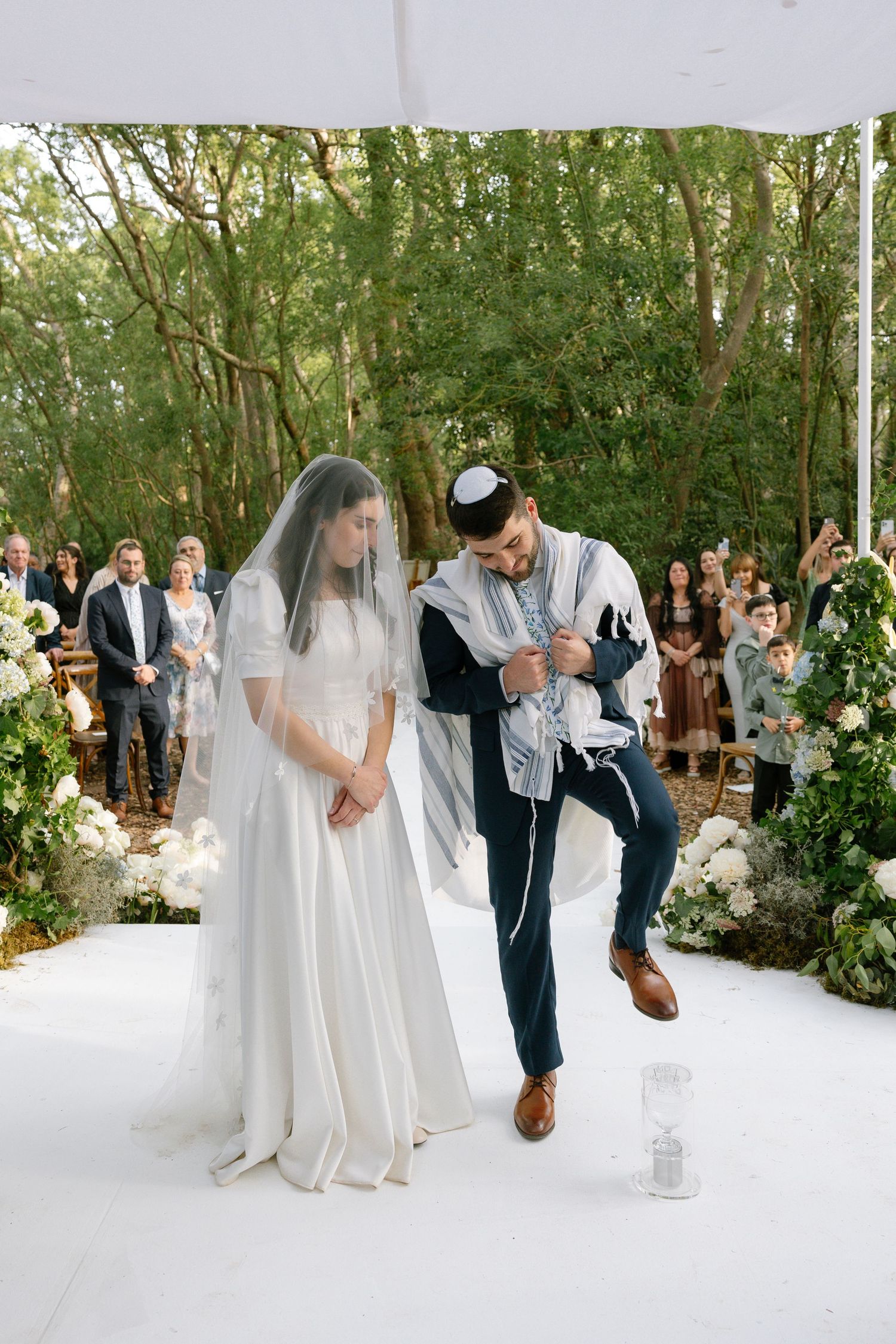 Wedding ceremony on white platform decorated with floral arrangements in a forest setting.