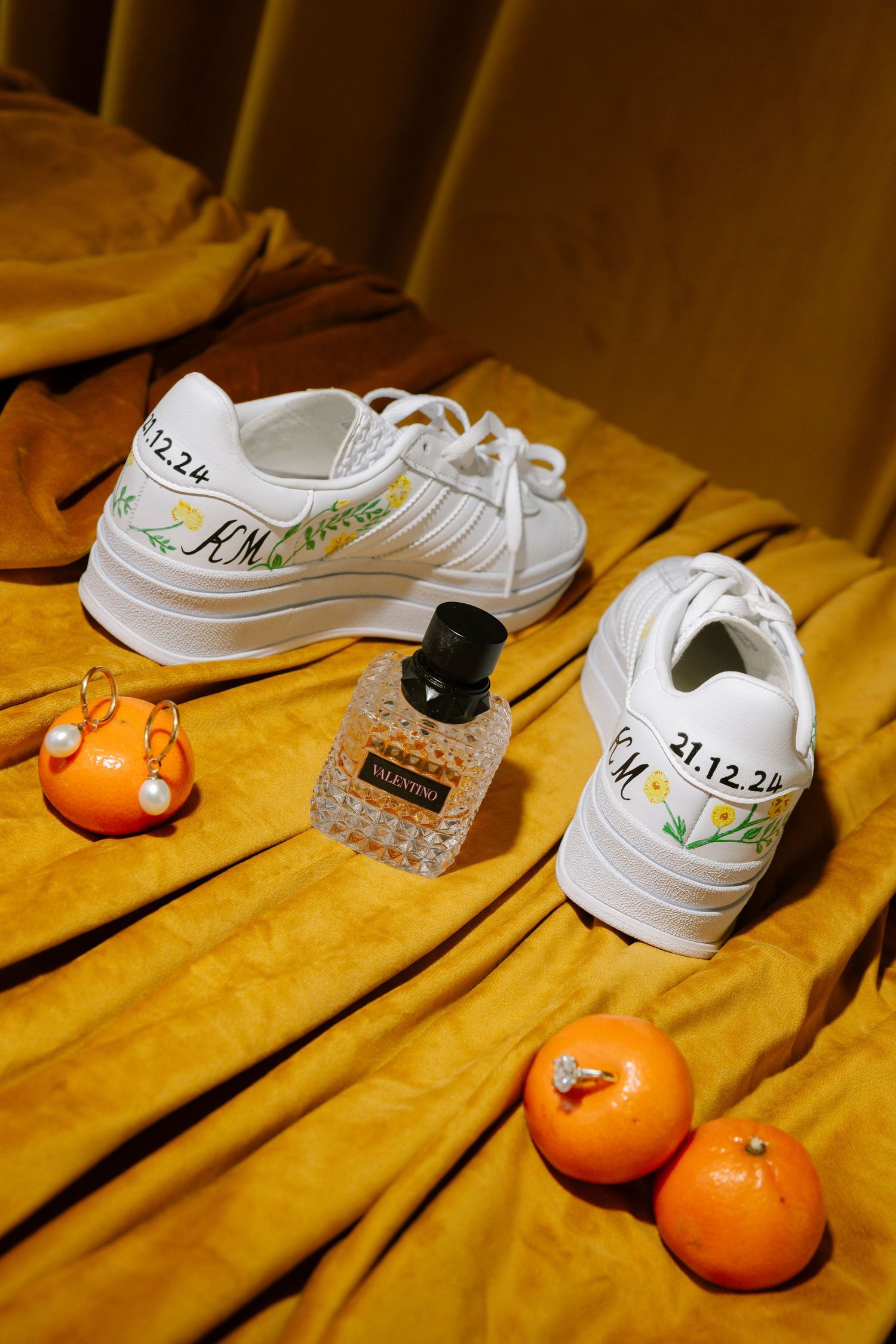 White sneakers arranged with oranges and small objects on a yellow velvet surface in a minimalist composition.