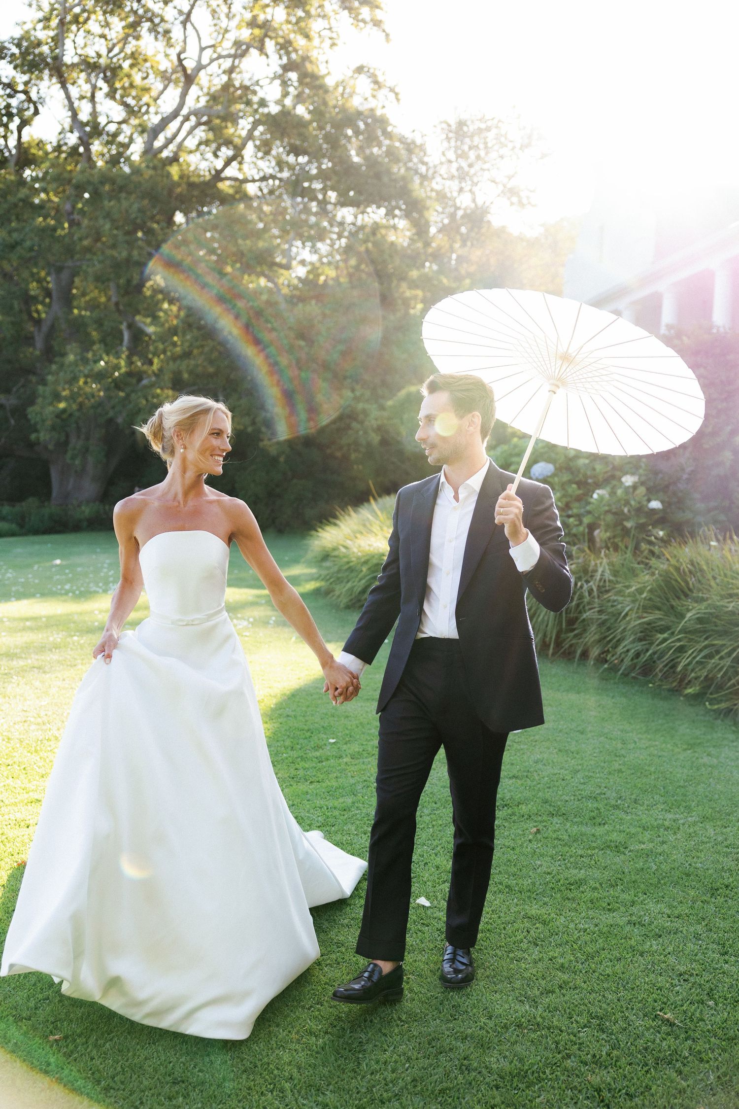 A couple strolls on a sunlit lawn, with the groom holding a white parasol as golden light streams through the trees.