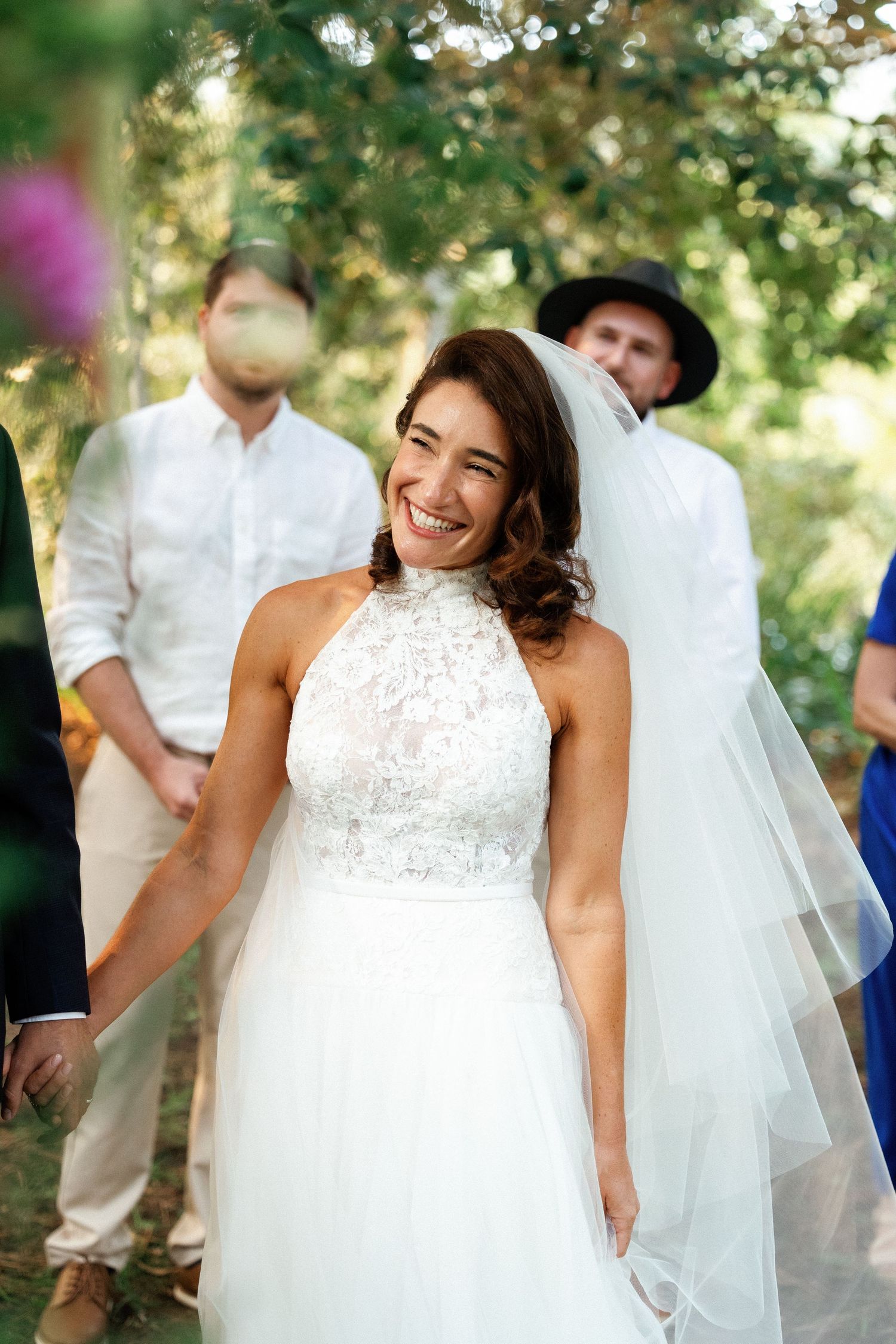 Smiling bride in white halter lace wedding dress and veil, standing outdoors among guests in a garden setting.