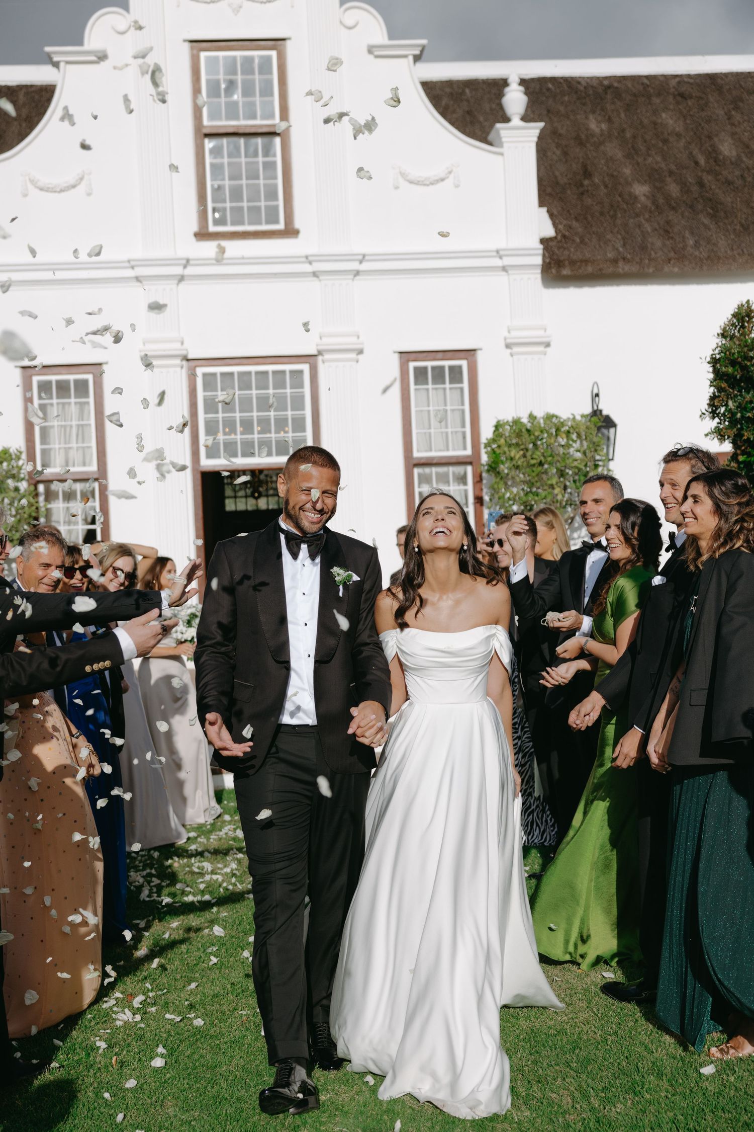 Wedding guests shower newlyweds with confetti on a grassy lawn outside a white colonial-style building.