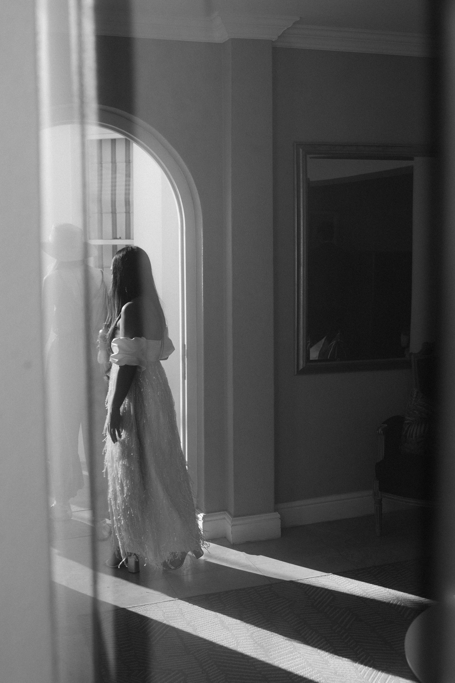 Black and white photo of a bride in a wedding dress and veil standing by an arched window in an elegant room.
