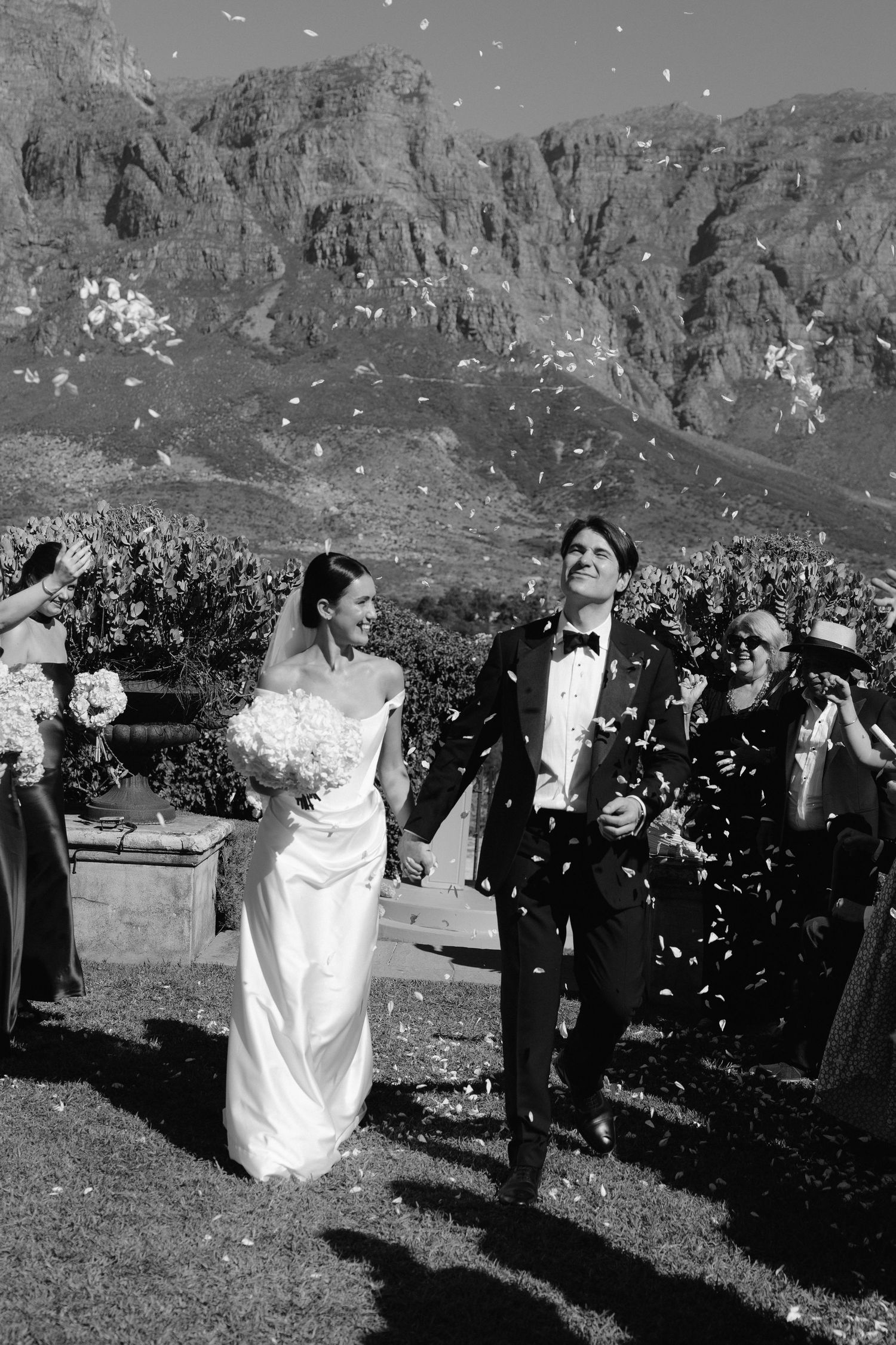 Black and white photo of a bride and groom walking through confetti with mountain backdrop at outdoor wedding.