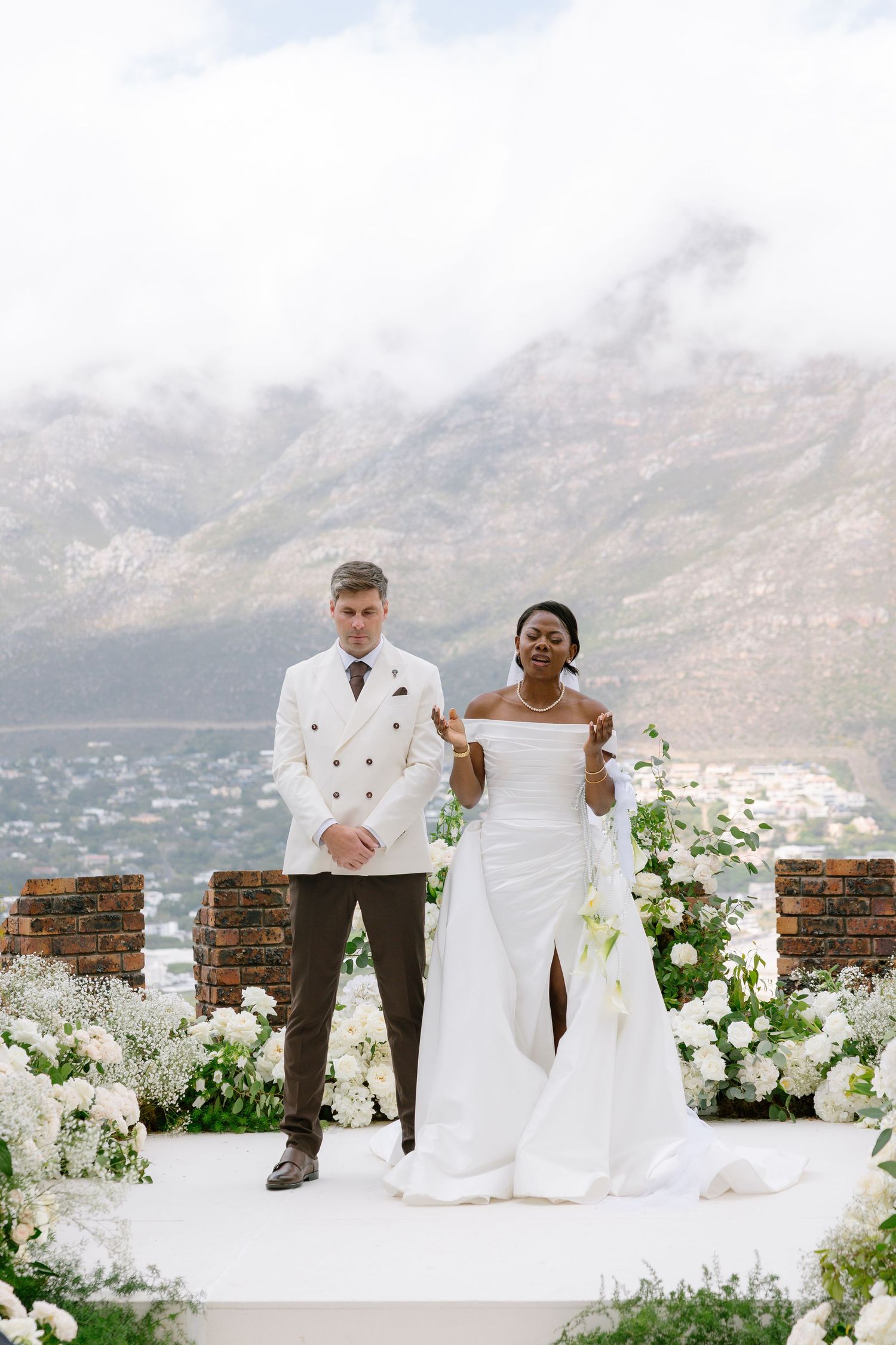 A wedding couple stands at a scenic mountain overlook decorated with white flowers and brick walls.