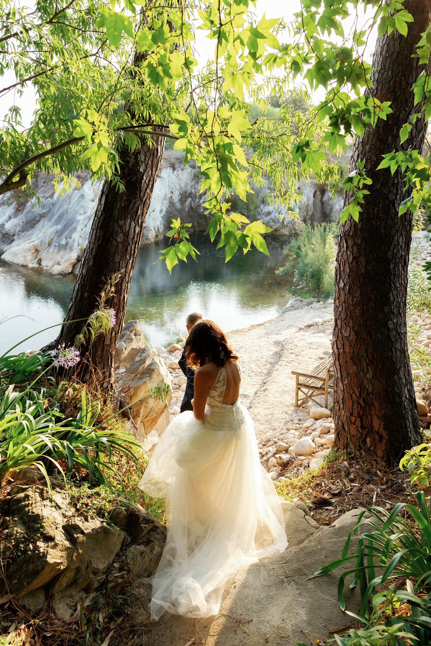 A figure in a flowing white dress walks along a sunlit forest path beside a tranquil stream.