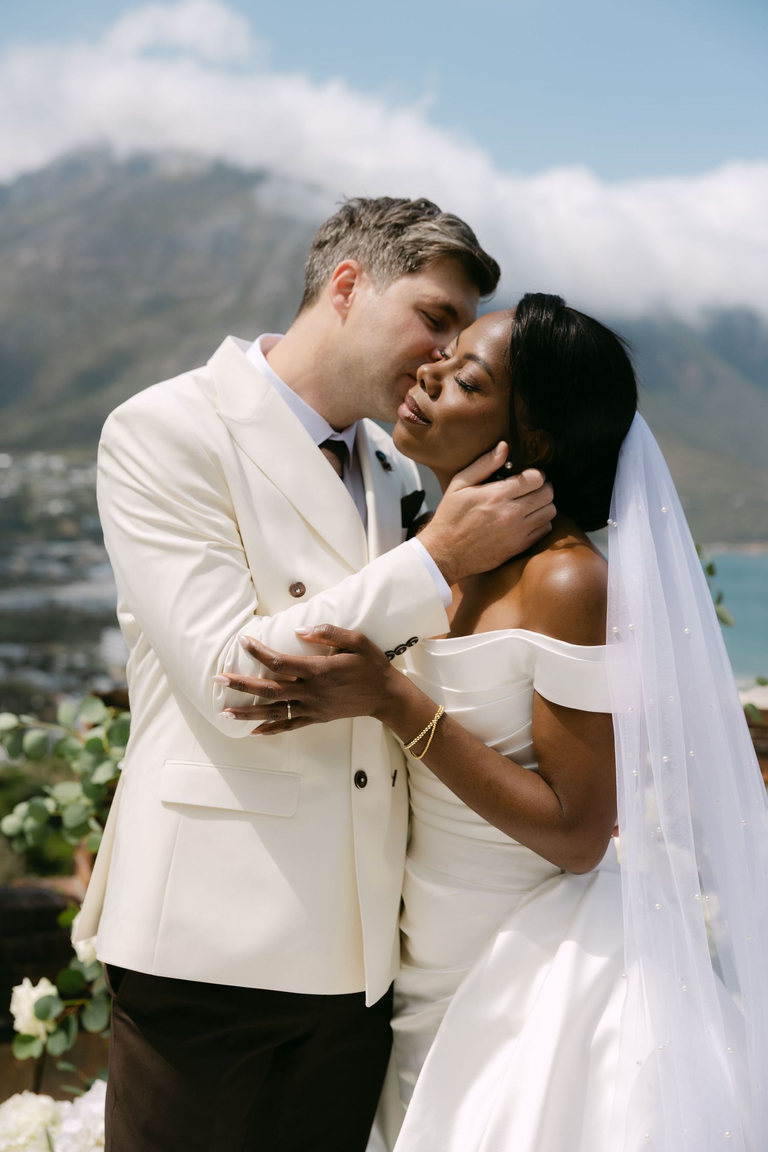 Interracial couple sharing a romantic kiss at their outdoor wedding with mountain backdrop.