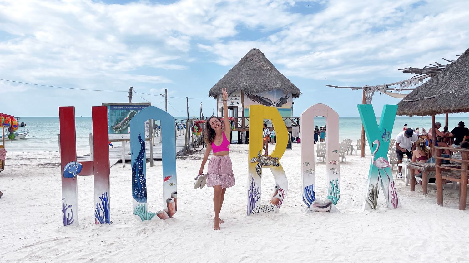Pink dress silhouette against large colorful 'HOLIDAY' beach sign with thatched hut in background.