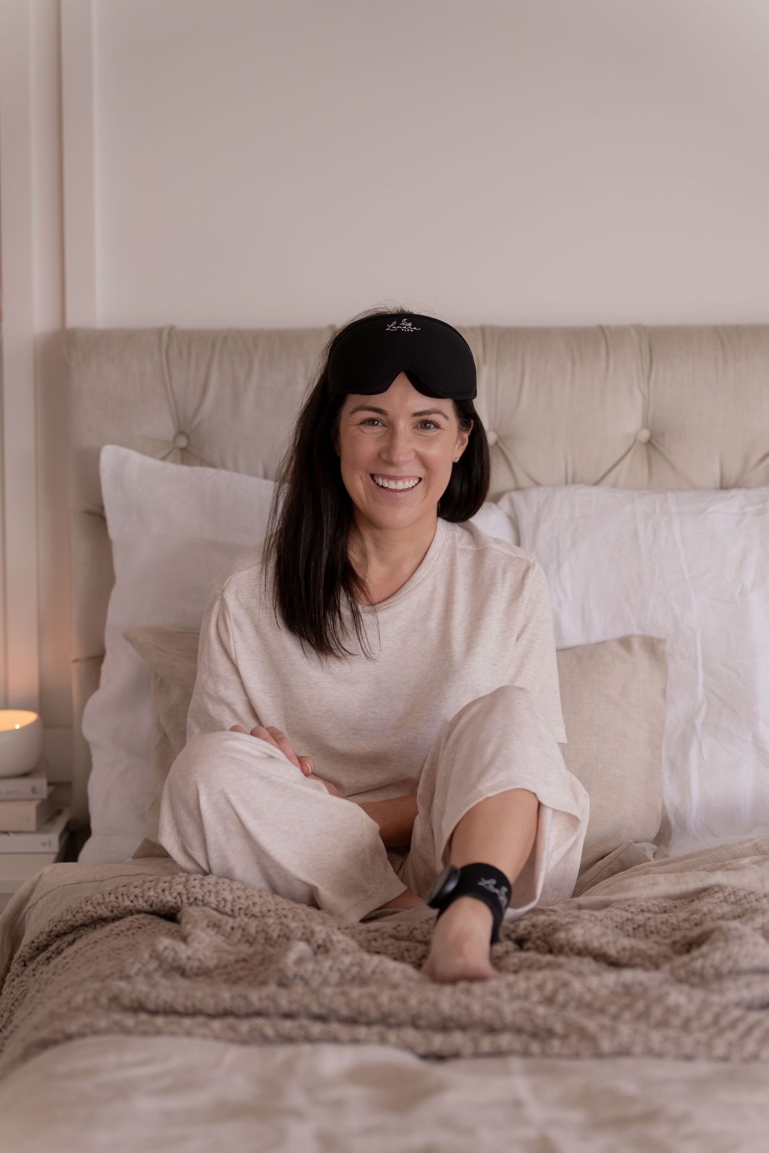 Woman in white pajamas and sleep mask smiling on a cozy bed with pillows and soft lighting.