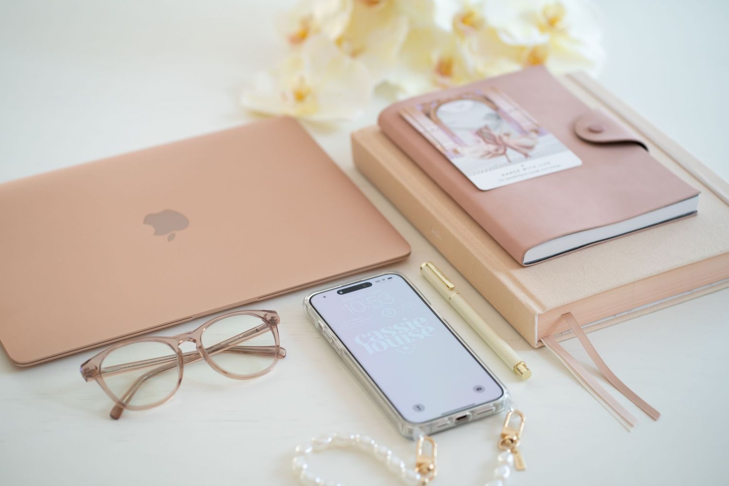 Rose gold laptop with glasses, phone and pink planner on white desk surface.