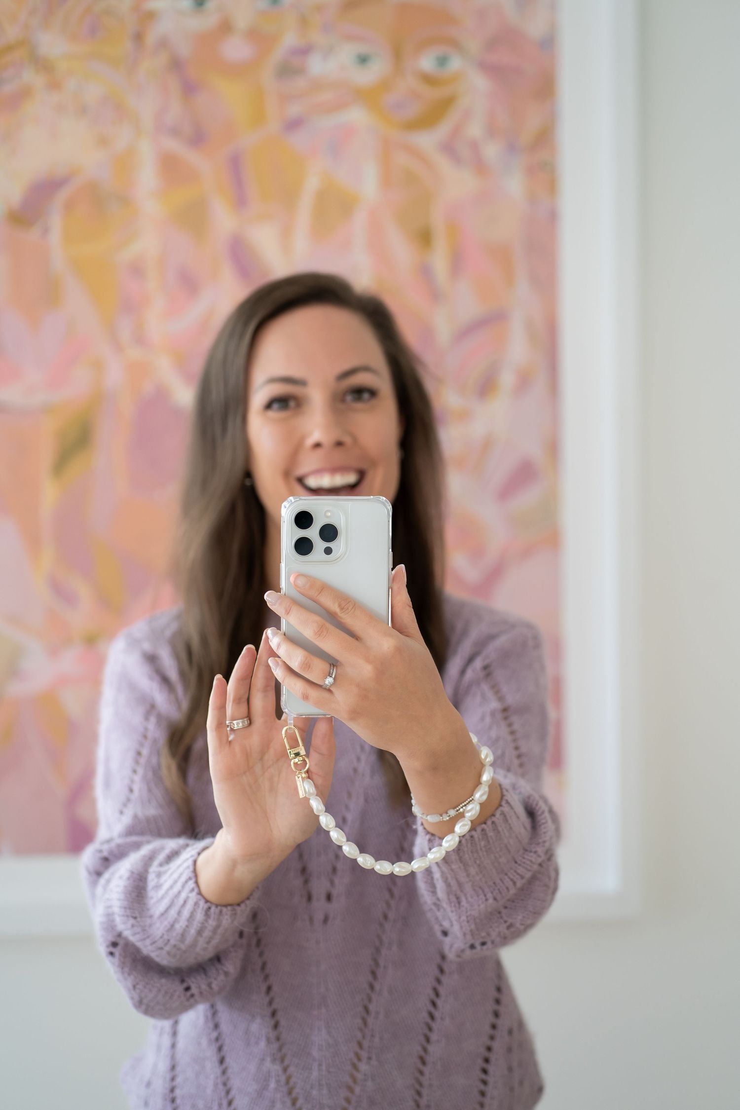 A person takes a mirror selfie while holding an iPhone, wearing a purple sweater against pink wall art.