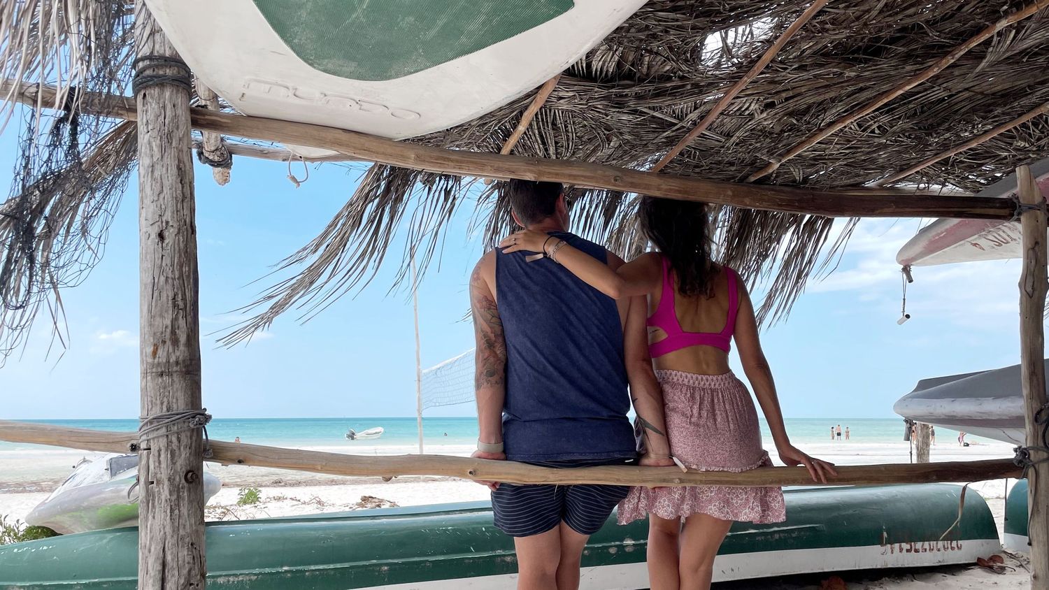 A couple enjoys a seaside view while standing under a thatched beach umbrella.