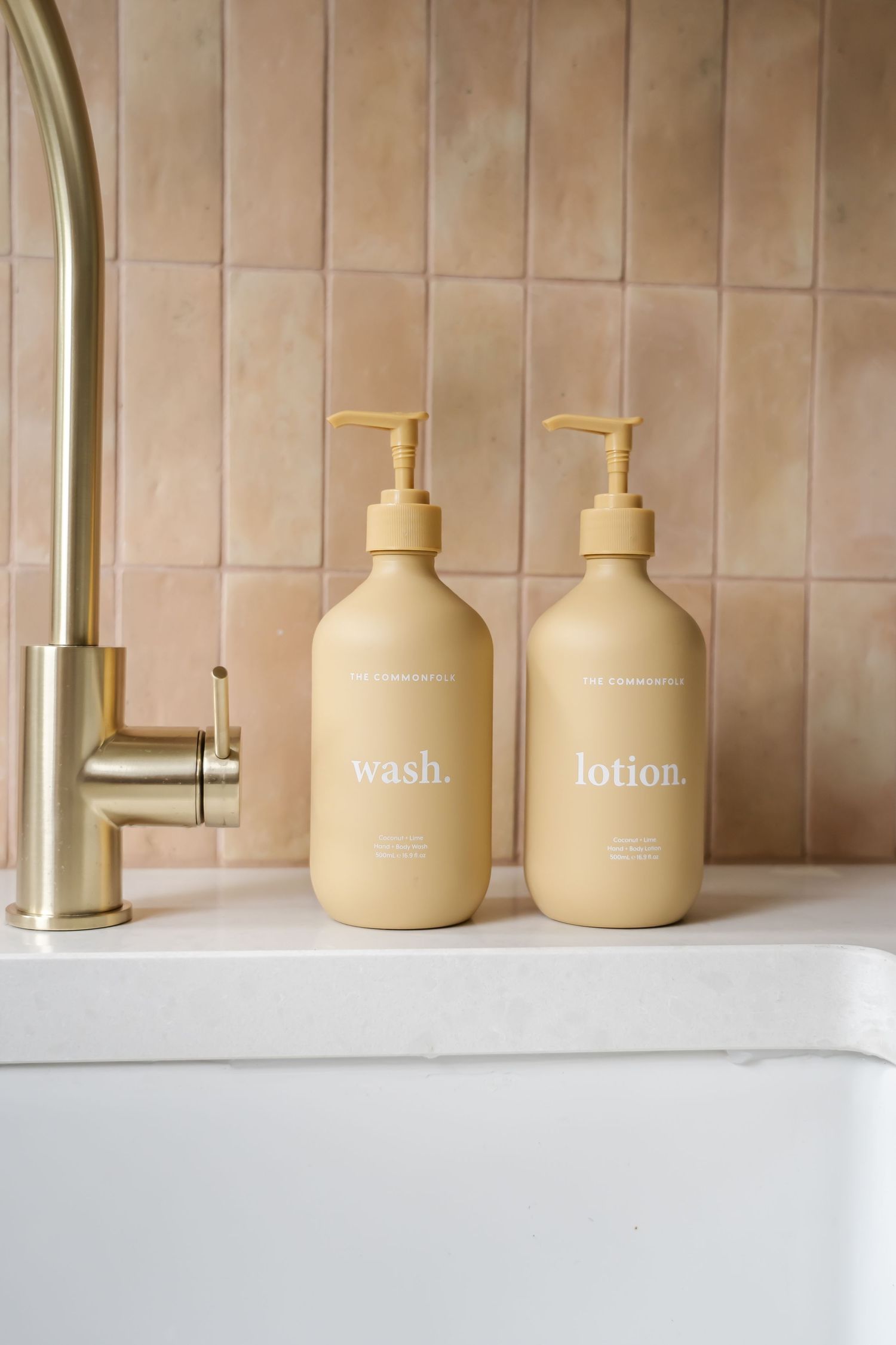 Two minimalist beige pump bottles labeled 'wash' and 'lotion' sit beside a gold faucet on a white counter.