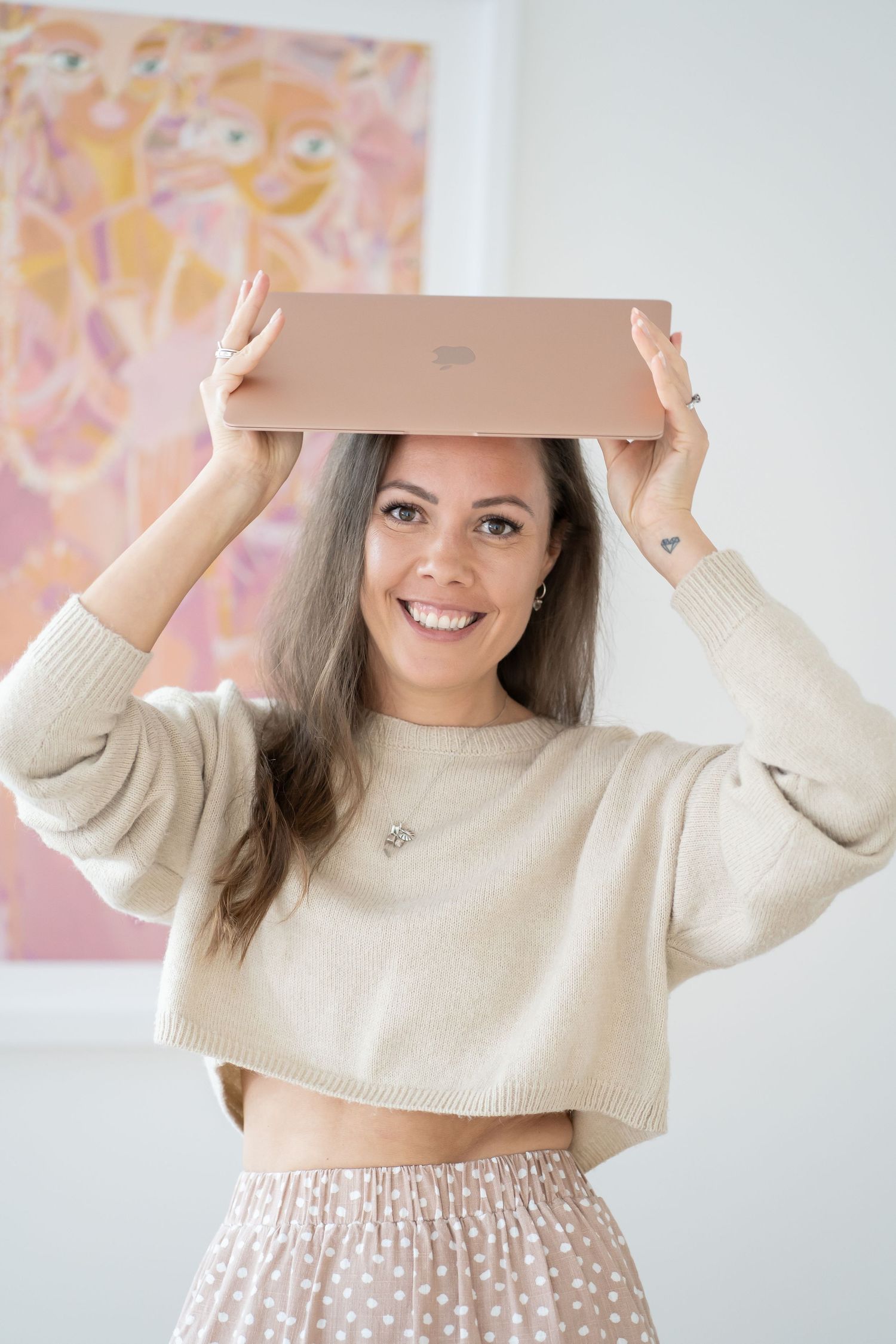 A person balances a rose gold laptop on their head while wearing a cream sweater and floral skirt.