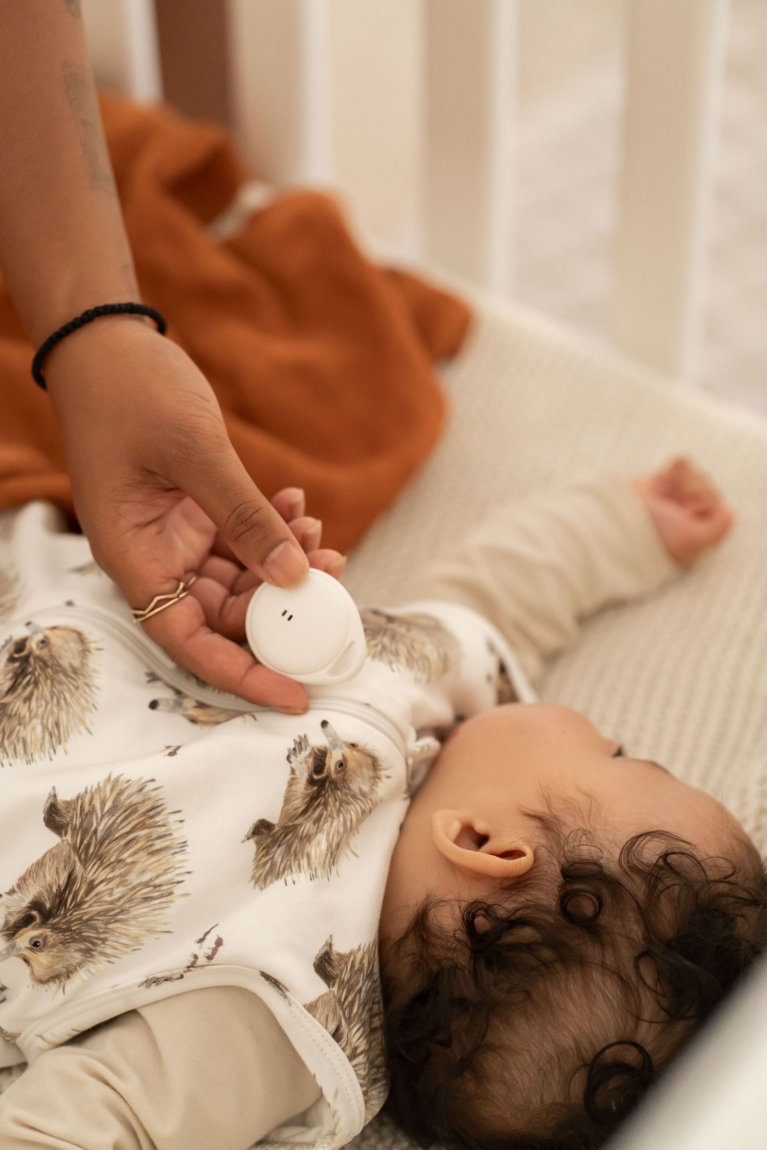 Baby lying in crib with white hedgehog-patterned bedding while a hand holds a small pacifier nearby.