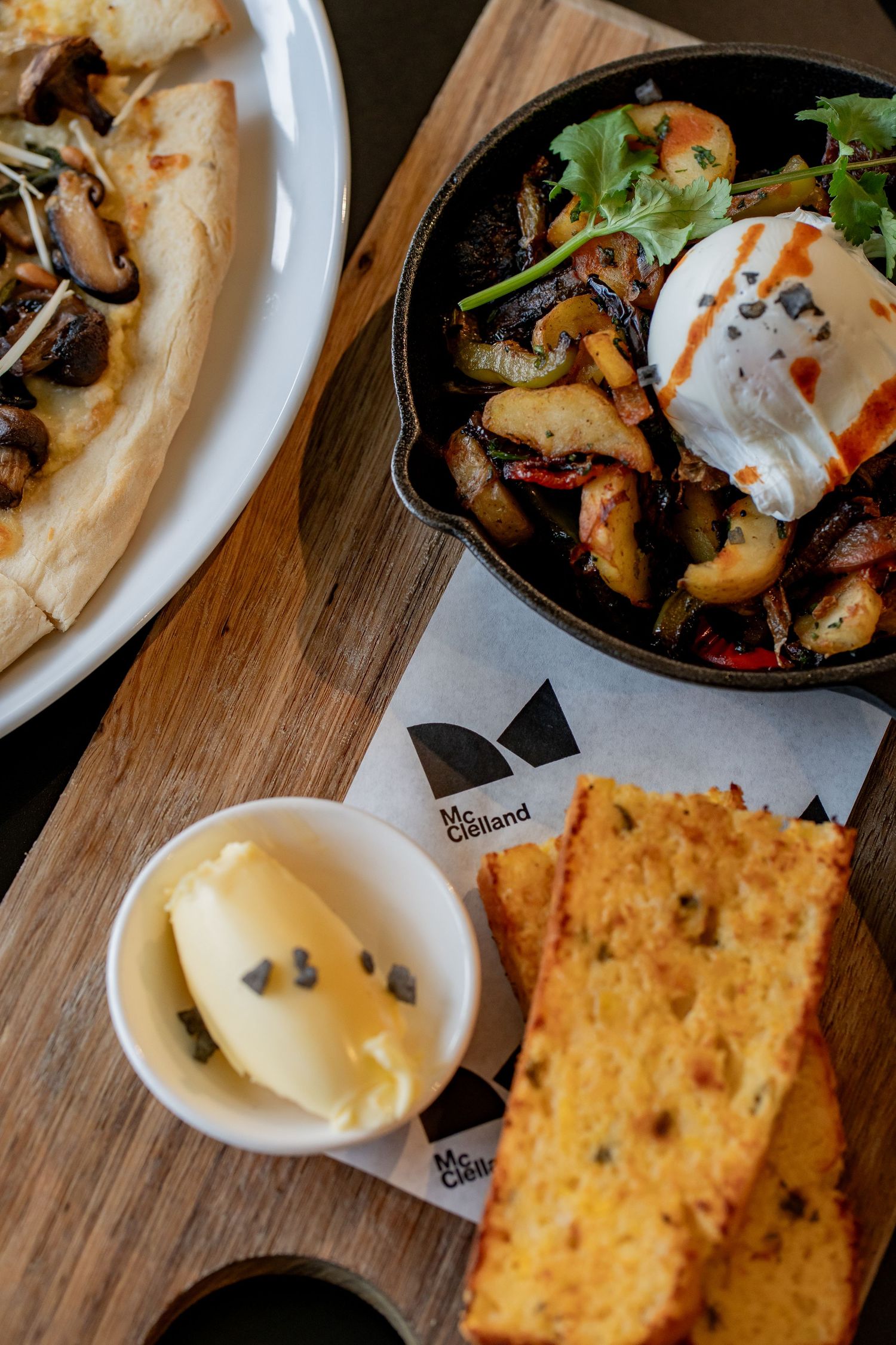 Rustic wooden board with garlic bread, butter, cast iron skillet with poached egg, and mushroom flatbread at McClelland cafe.