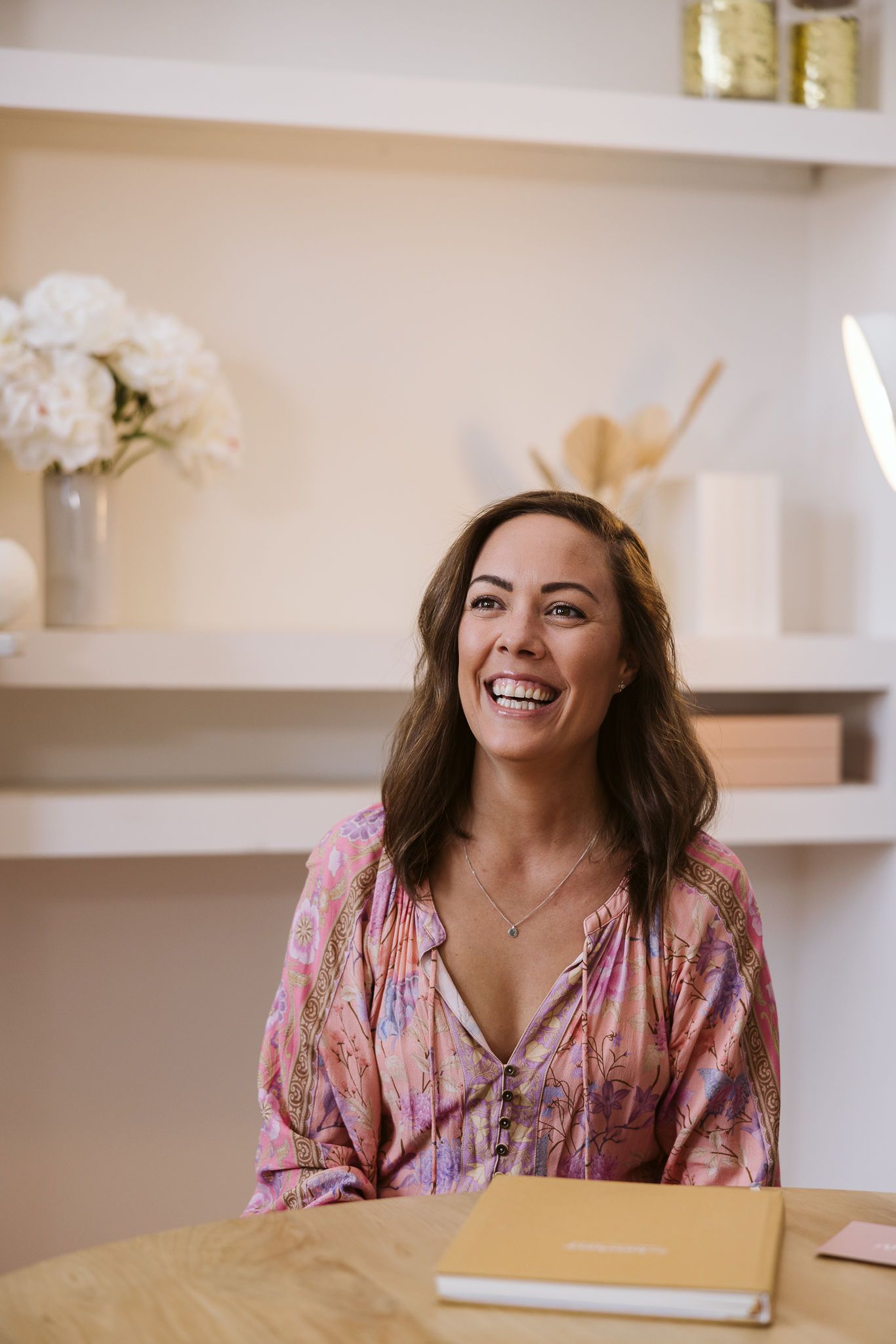 Woman in pink floral top smiling while sitting at desk with white shelving and floral decor in background.