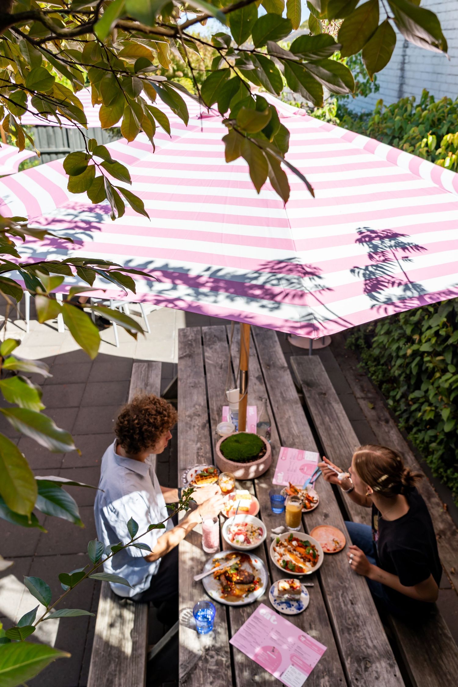 A summertime outdoor meal under a pink and white striped umbrella on a wooden picnic table with tree leaves overhead.