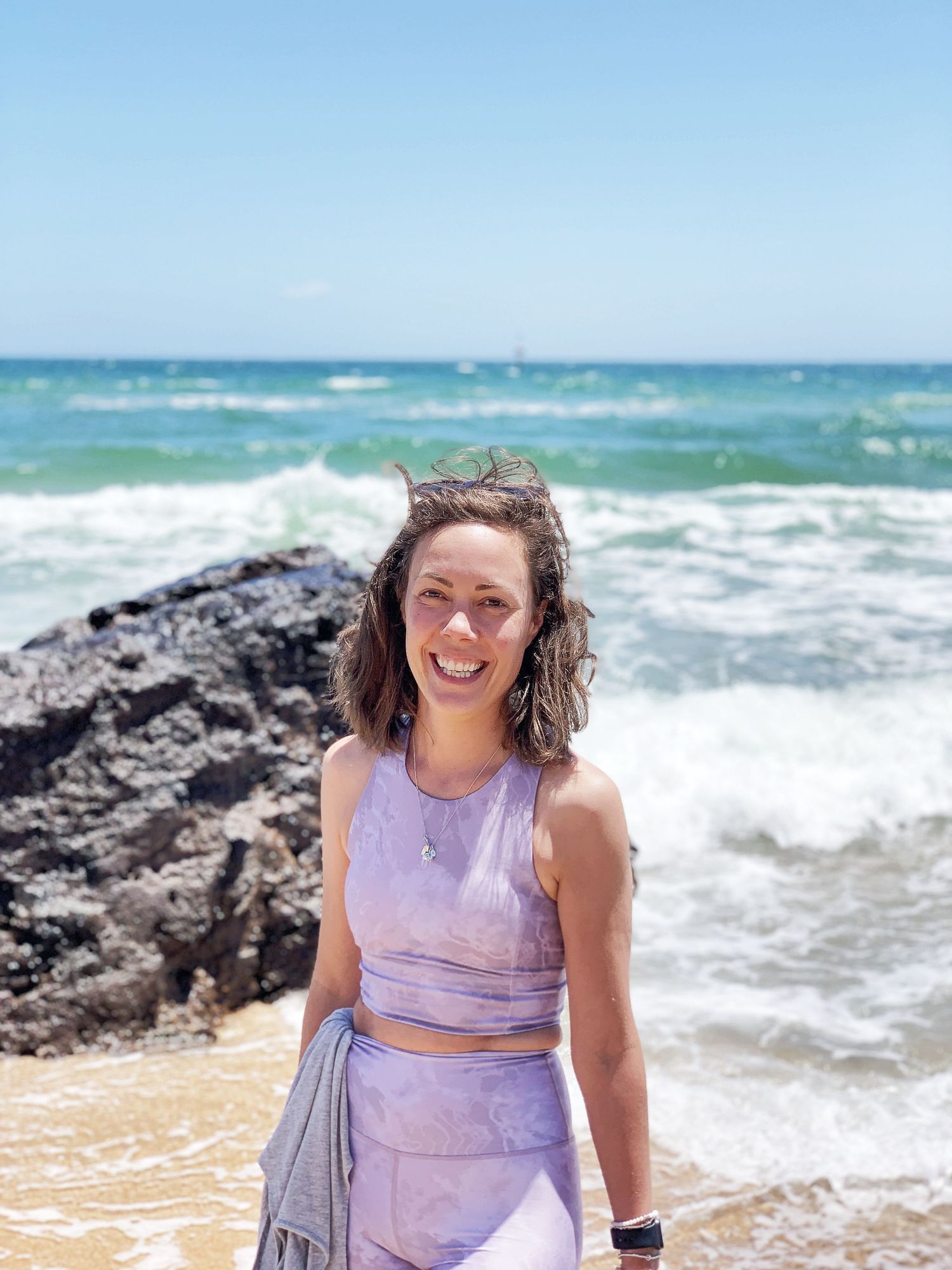 A person in lavender workout clothes poses happily beside crashing waves and black rocks at the beach.