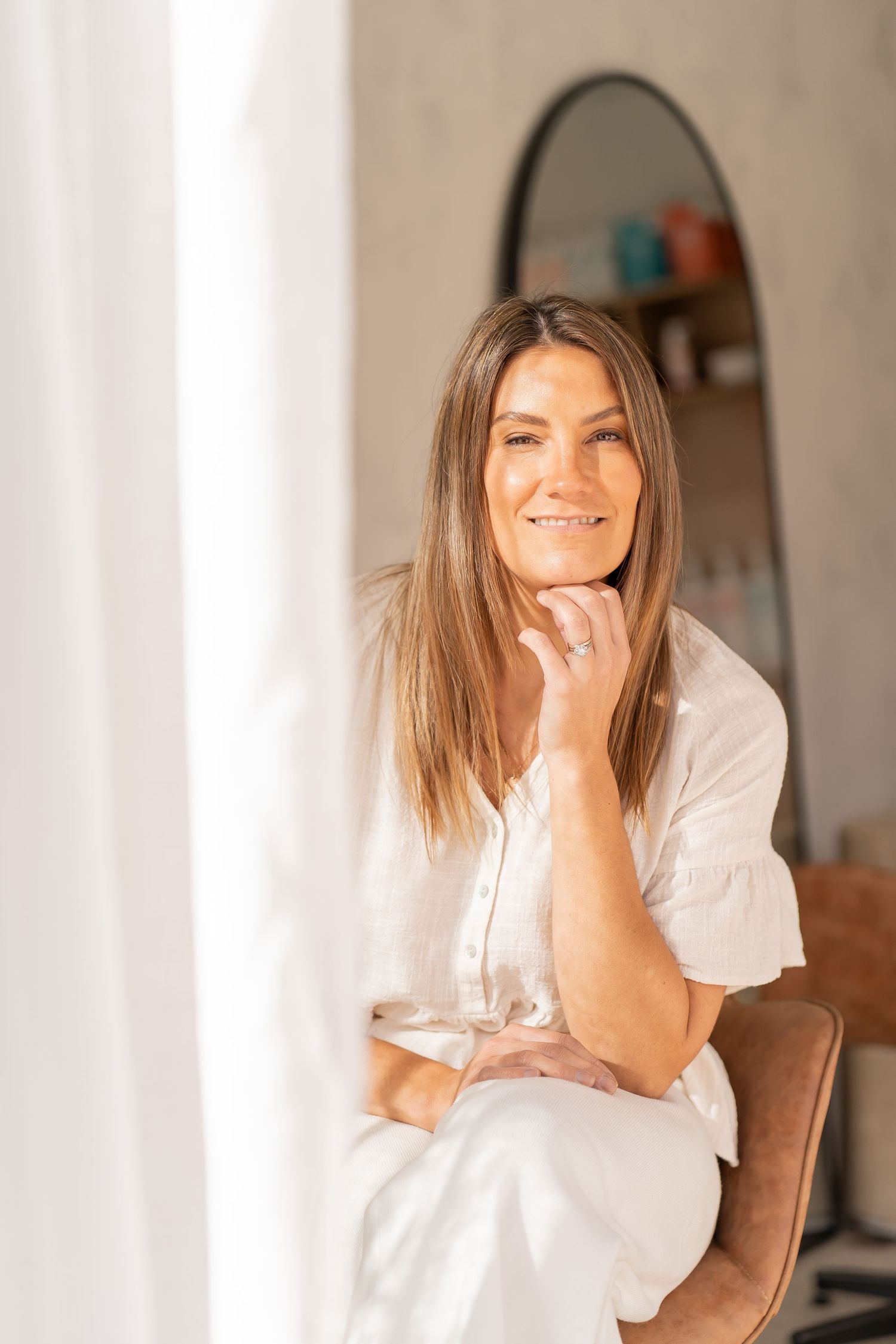 A person in a white shirt sits on a couch smiling warmly while resting their chin on their hand.