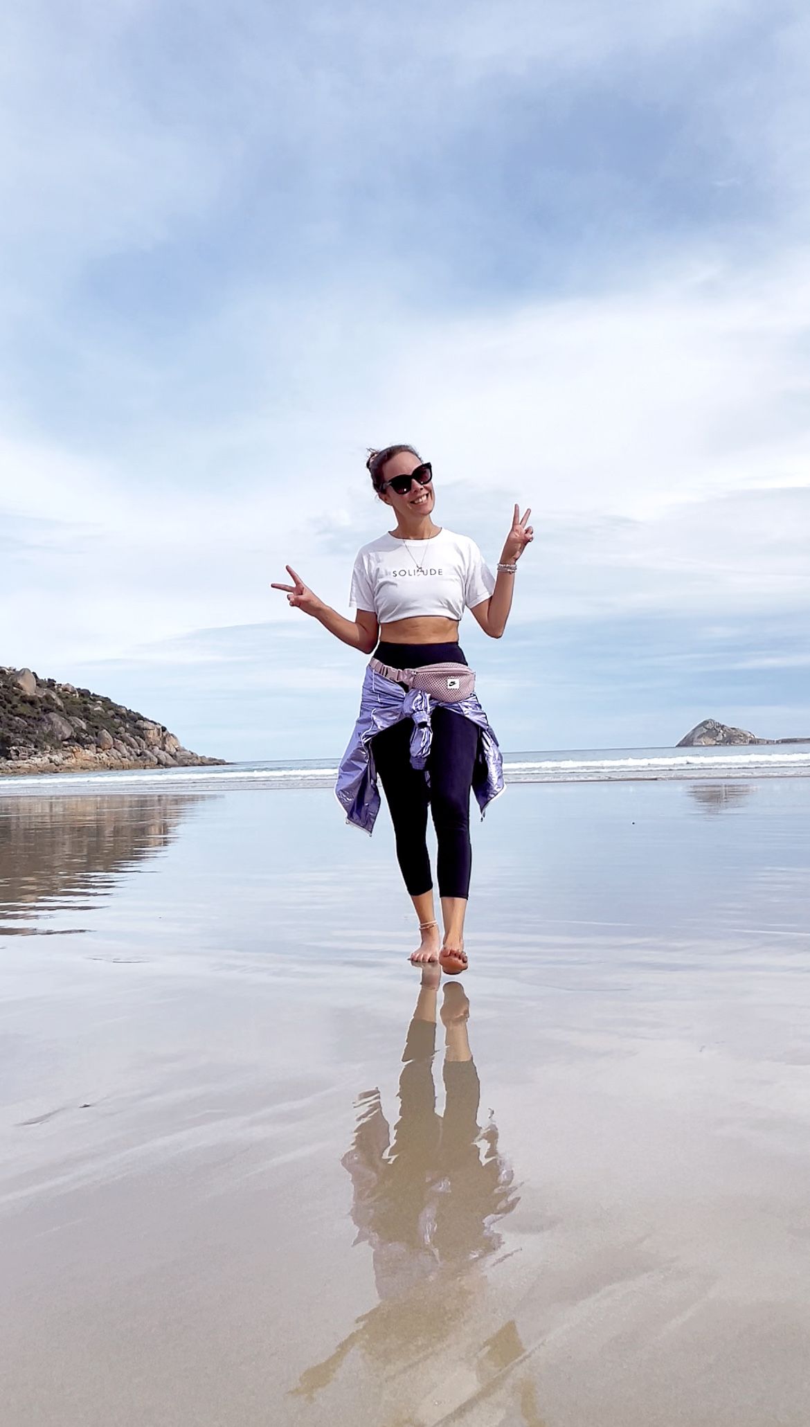 A person in athletic wear makes peace signs while jumping on a beach with their reflection visible in wet sand.