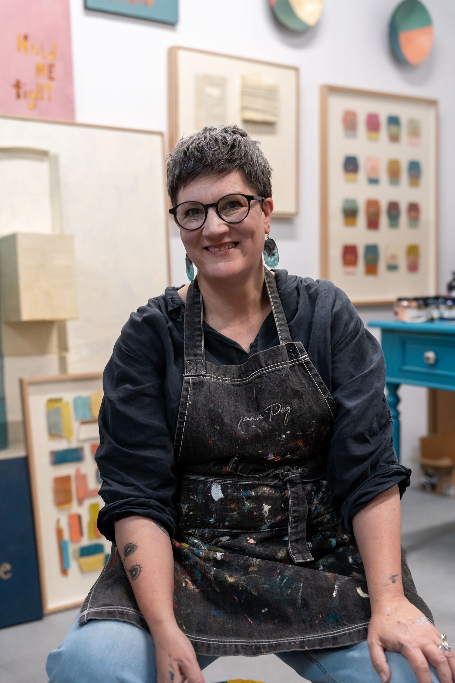 Smiling female artist in paint-splattered denim apron sits in her colorful art studio surrounded by framed artwork.