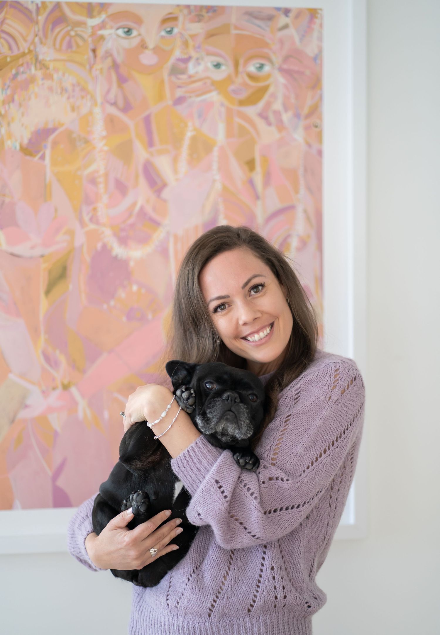 A person in a purple sweater holds a small black pug dog while standing in front of pink abstract artwork.