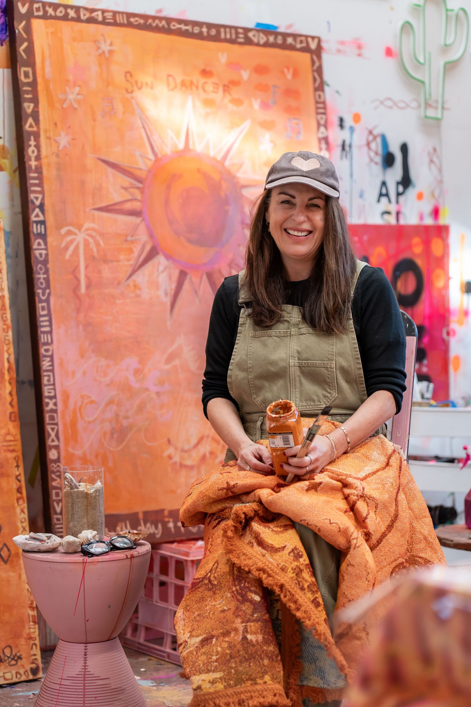 Smiling female artist in olive apron and cap sits in colorful studio holding ceramic piece before orange sun painting.