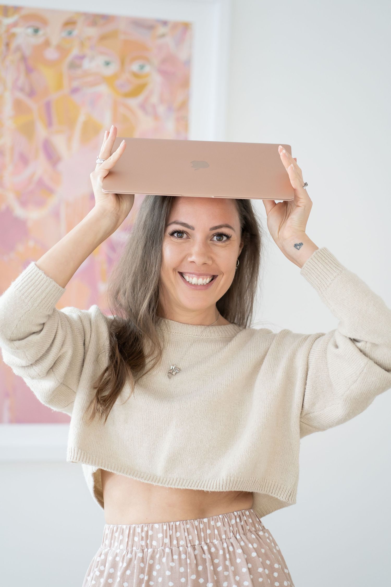 Smiling woman balancing a rose gold Apple MacBook on her head, wearing a beige crop sweater and polka dot skirt.