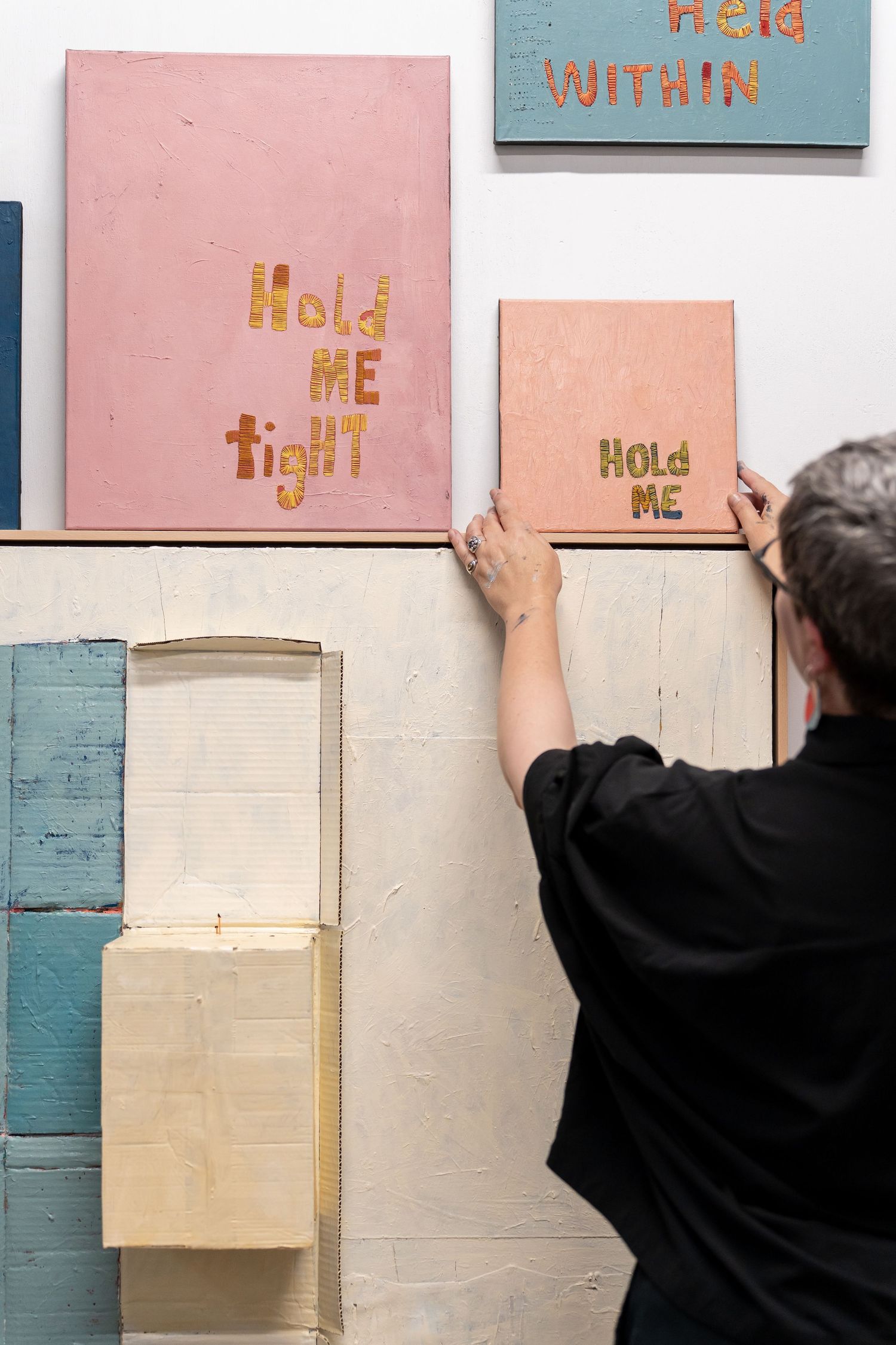 Artist arranging pink canvas artworks with gold text reading Hold Me Tight and Hold Me on a white gallery wall.