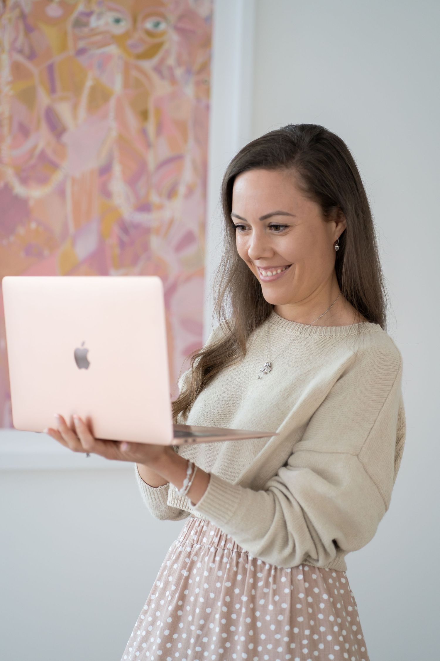 Someone in a cream sweater and polka dot skirt smiles while working on a rose gold MacBook against a pink wall.