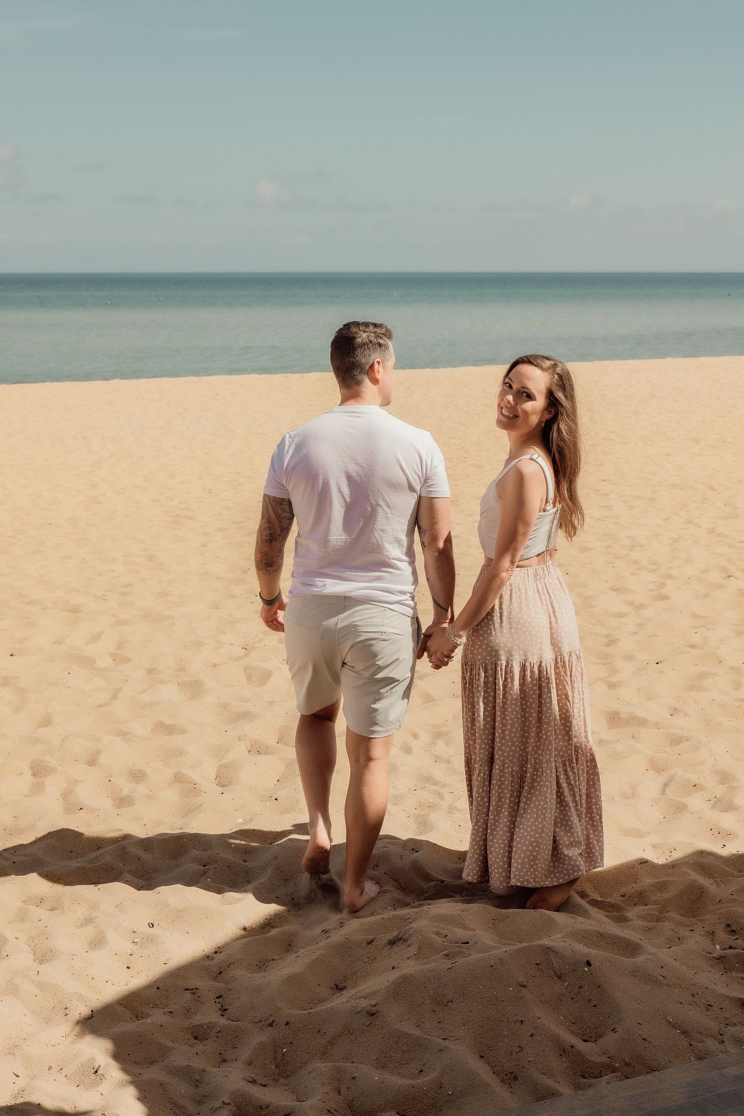 Couple holding hands while walking on sandy beach with ocean view in the background.