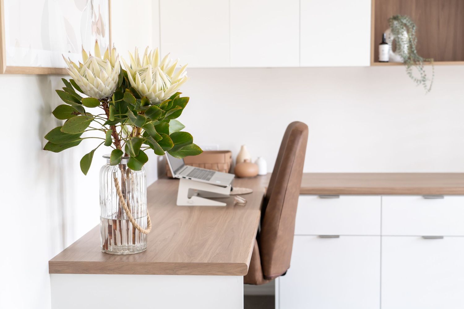 Modern home office desk with white protea flowers in glass vase, wooden desktop, and brown chair.