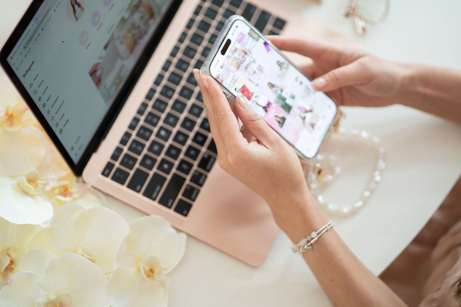 Hands holding smartphone showing social media app next to MacBook on floral desk setup.