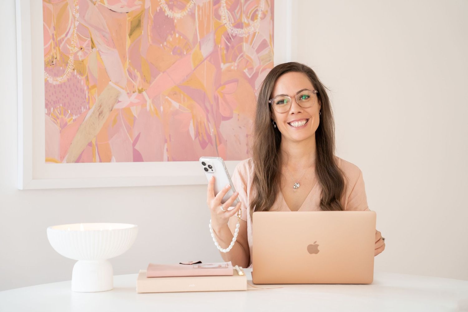 A person sits at a clean white desk with laptop and phone, wearing glasses in front of pink artwork.