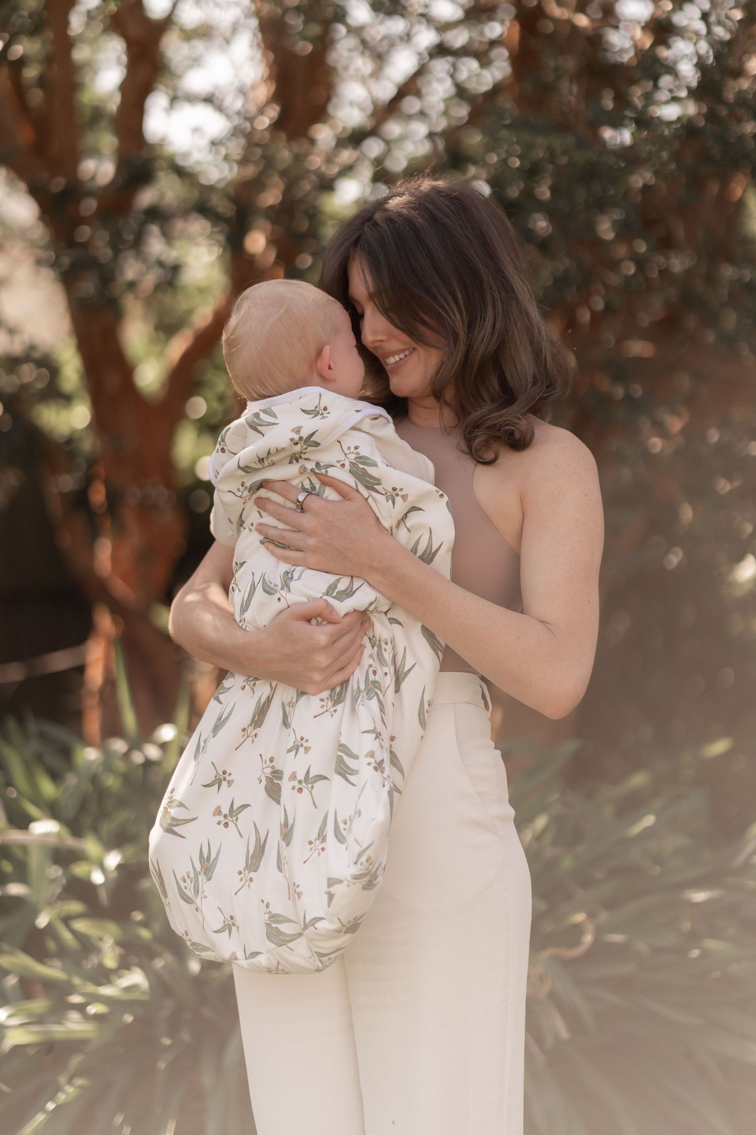 Embracing family bond in nature with baby wearing a floral swaddle and sunlit trees in background.