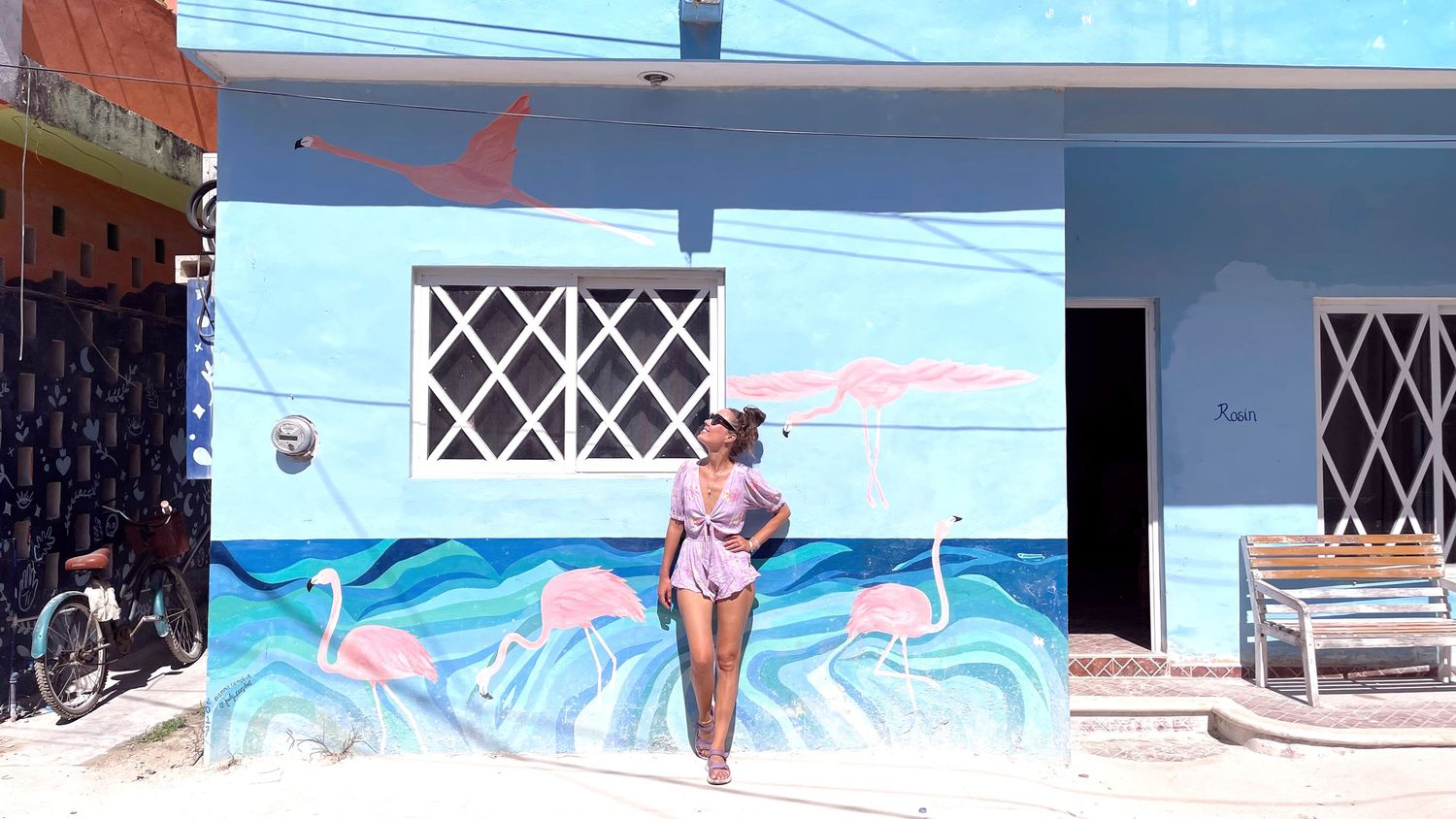 Person in pink outfit standing against colorful wave mural on blue wall near beach.
