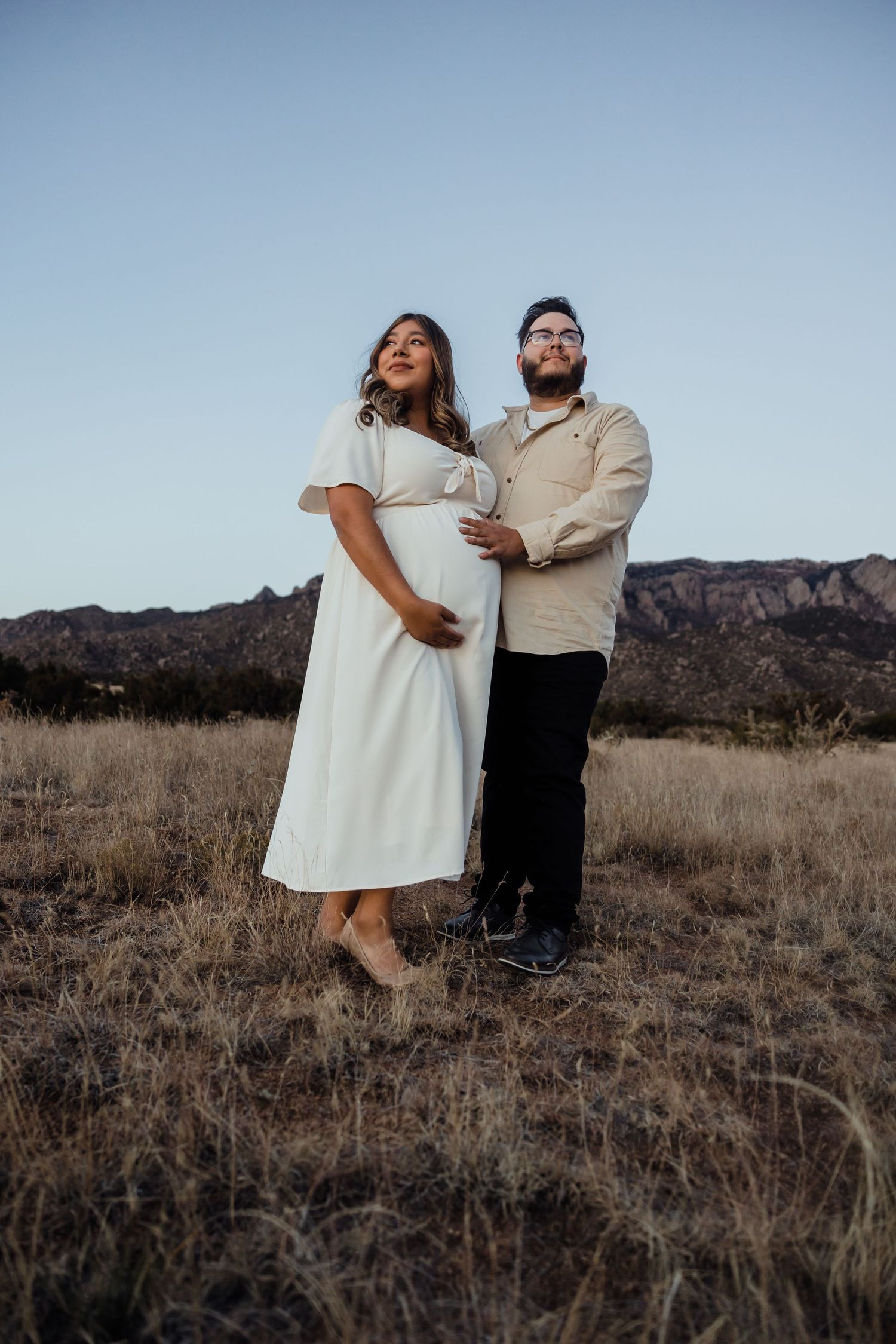 Pregnant couple posing outdoors in a desert field at dusk, woman in white dress cradling baby bump, mountains in background.