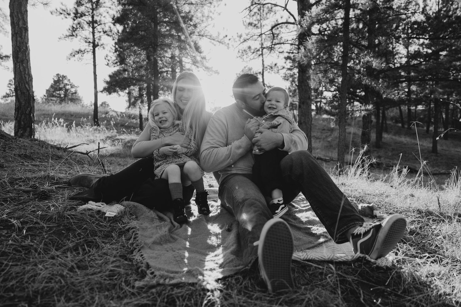 Black and white photo of a happy family sitting together on grass in a sunlit forest setting.