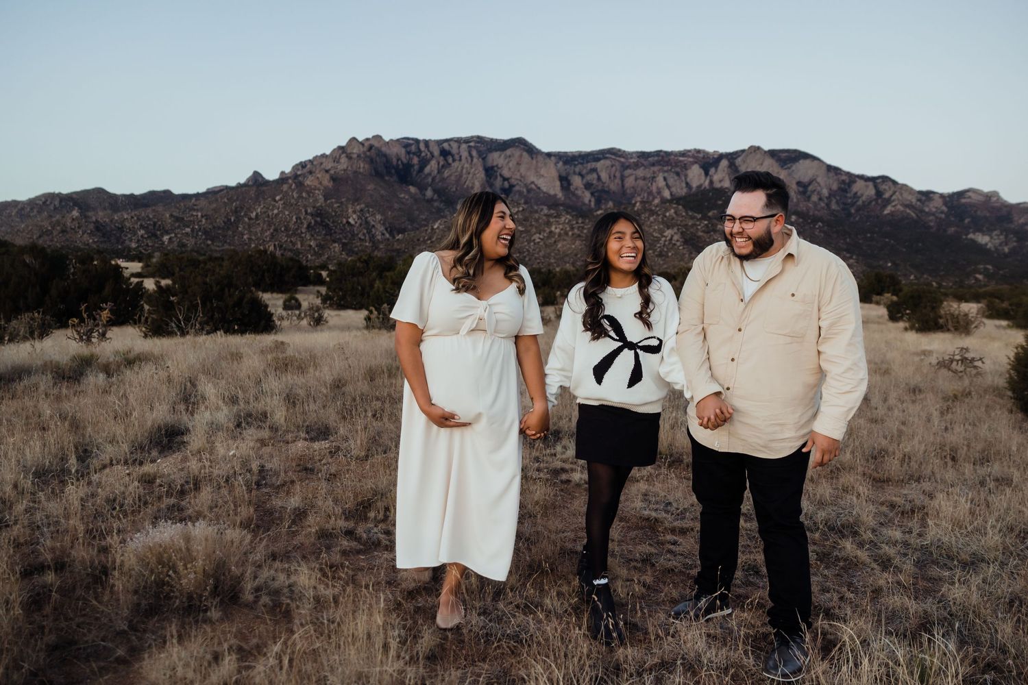 Pregnant woman in white dress with two companions in a field with Sandia Mountains backdrop.