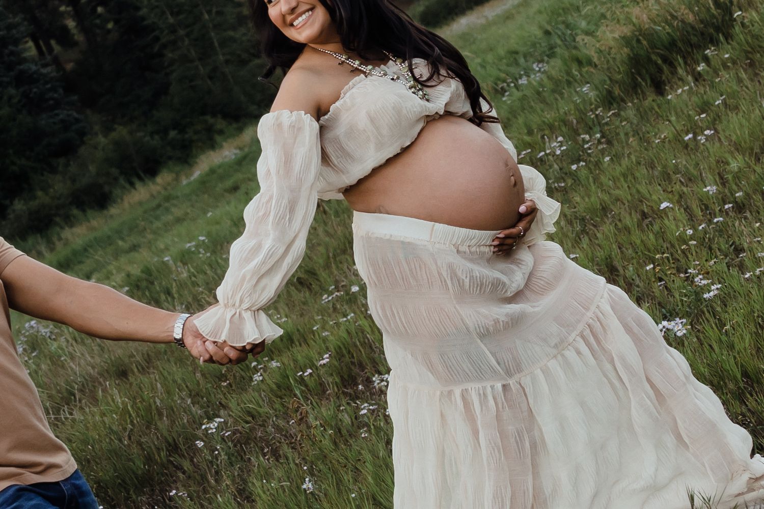 Pregnant woman in white off-shoulder dress holding hands with partner in a wildflower meadow during maternity photoshoot.