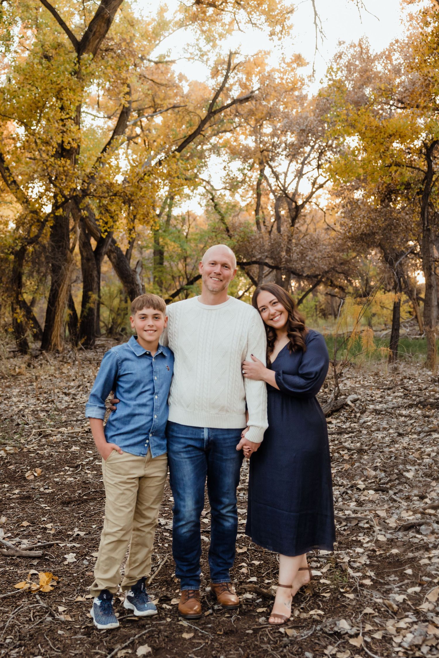 Family of three posing outdoors in autumn forest with colorful yellow foliage, wearing casual blue and navy outfits.